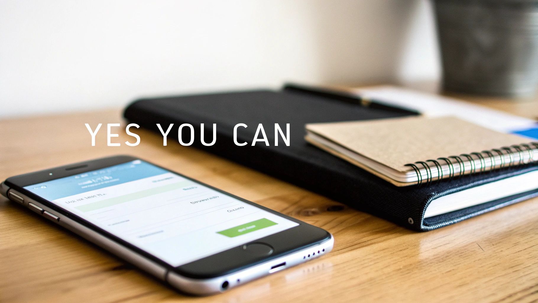 A smartphone and notebooks on a wooden desk with the text "YES YOU CAN", suggesting a motivated workspace.