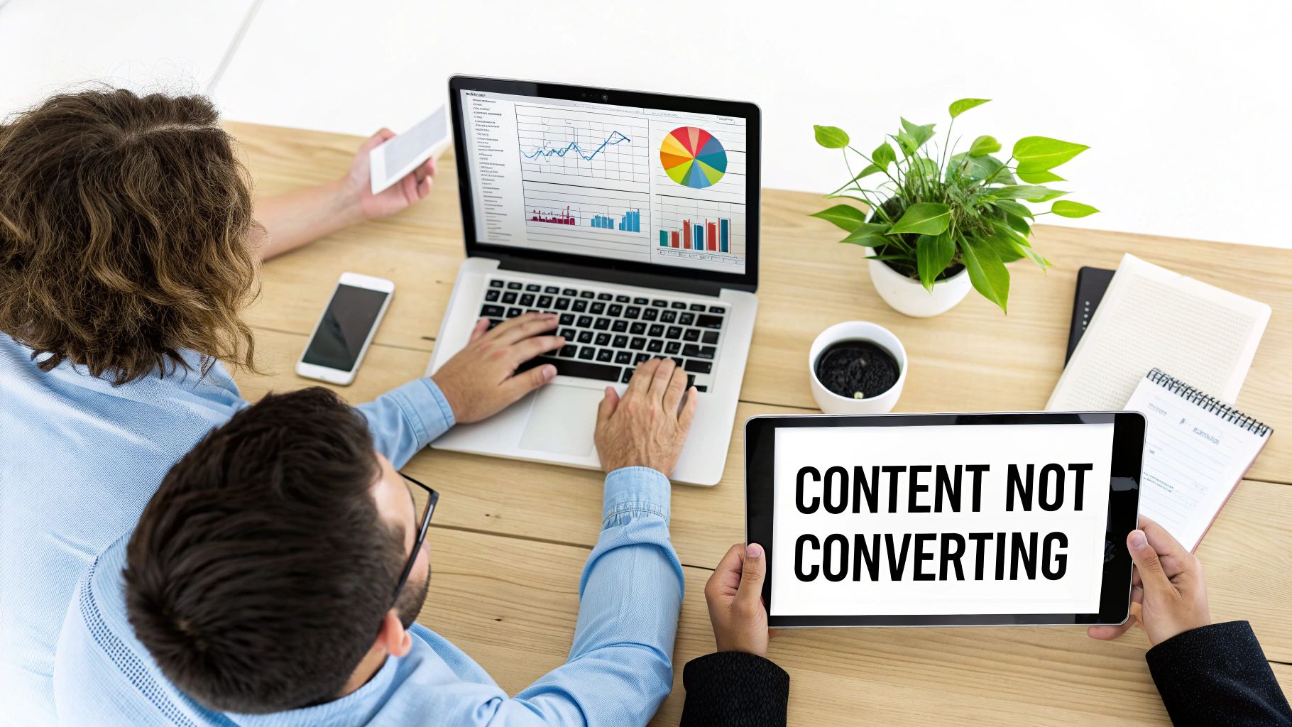 Overhead view of two marketers analyzing data on a laptop and discussing content conversion issues on a tablet.