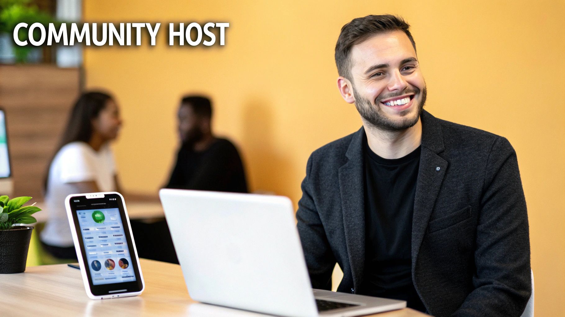 A smiling man, possibly a community host, sits at a desk with a laptop and a phone.