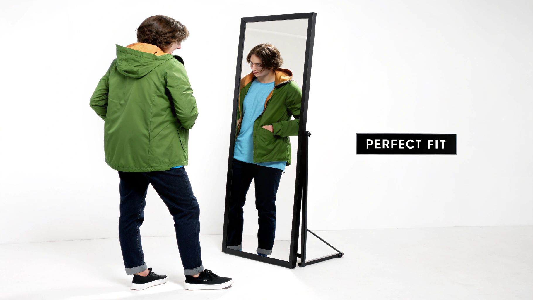A young man wearing a green jacket and blue jeans looks at his reflection in a mirror, showing a perfect fit.