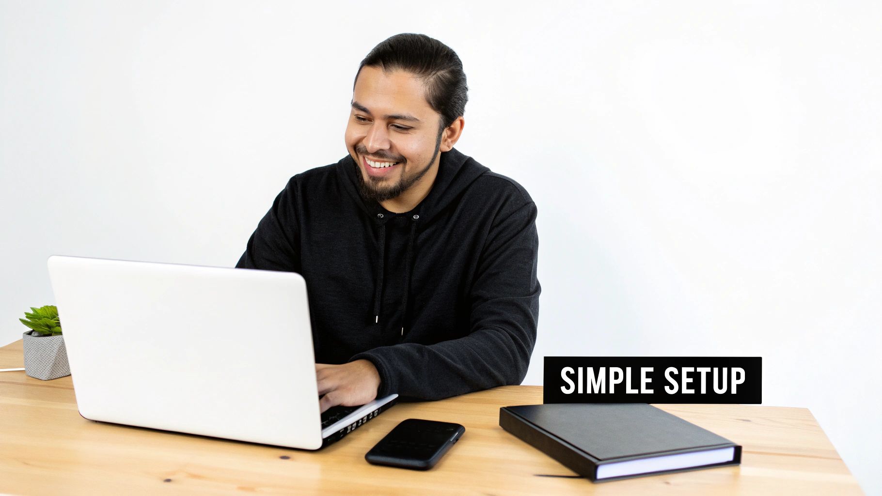 A smiling man in a black hoodie works on a laptop at a simple wooden desk setup.