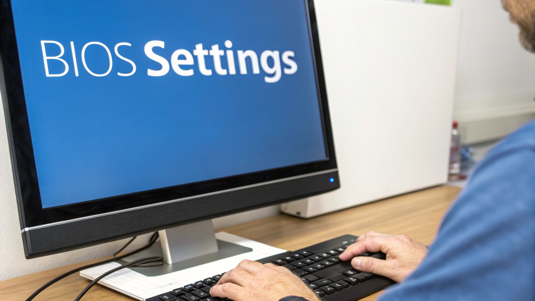 A man types on a keyboard, facing a computer monitor displaying 'BIOS Settings' on a blue screen.