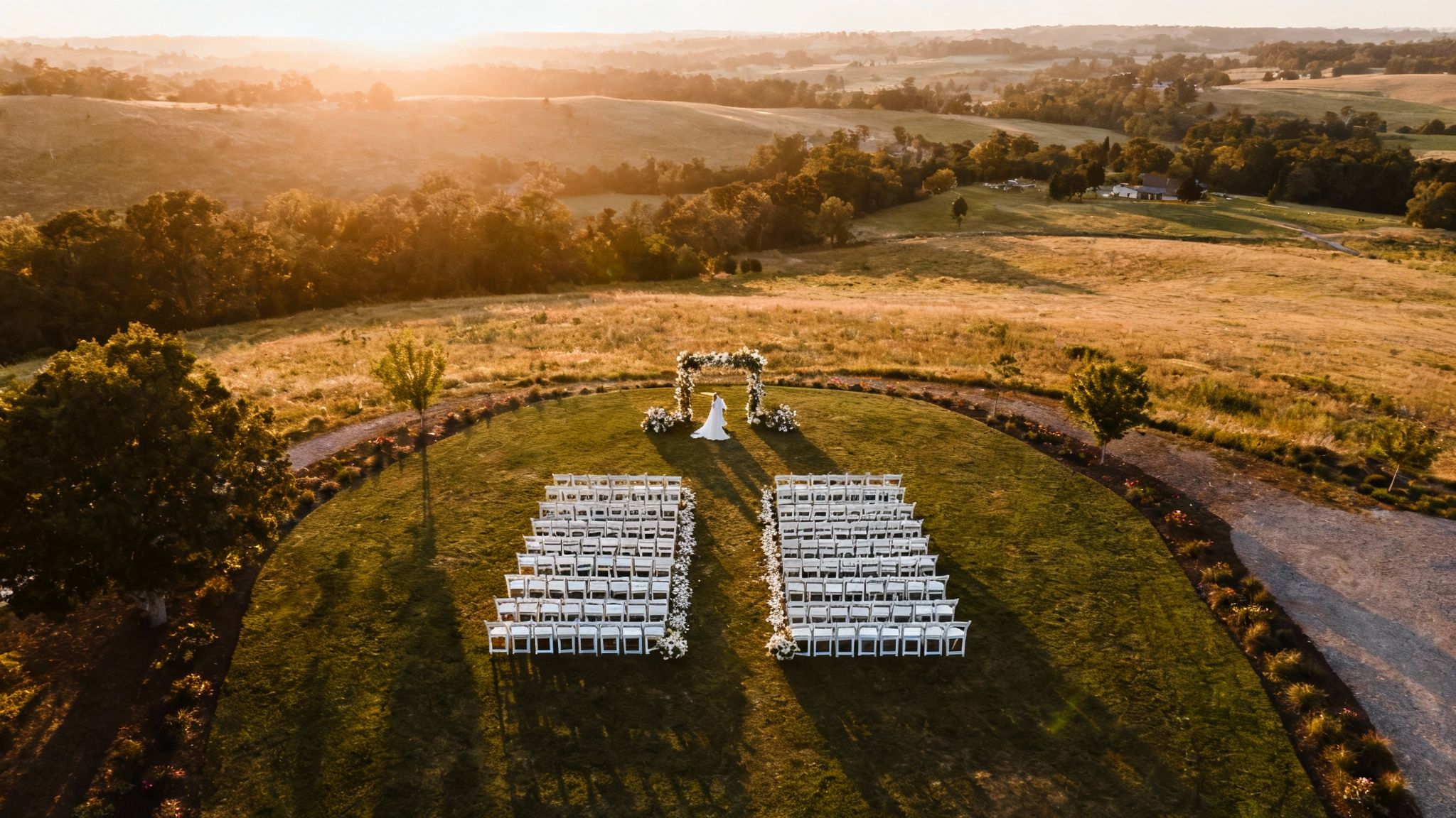 Aerial view of a golden hour outdoor wedding ceremony with a bride and white chairs.