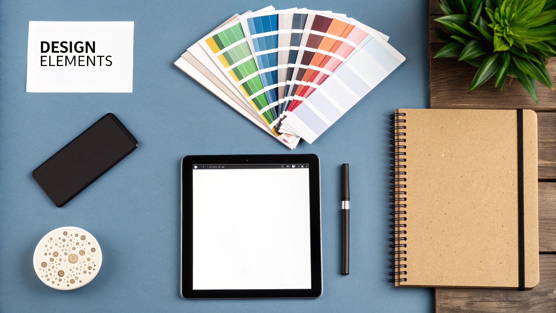 A flat lay of design elements including color swatches, a tablet, phone, and notebook on a blue desk.