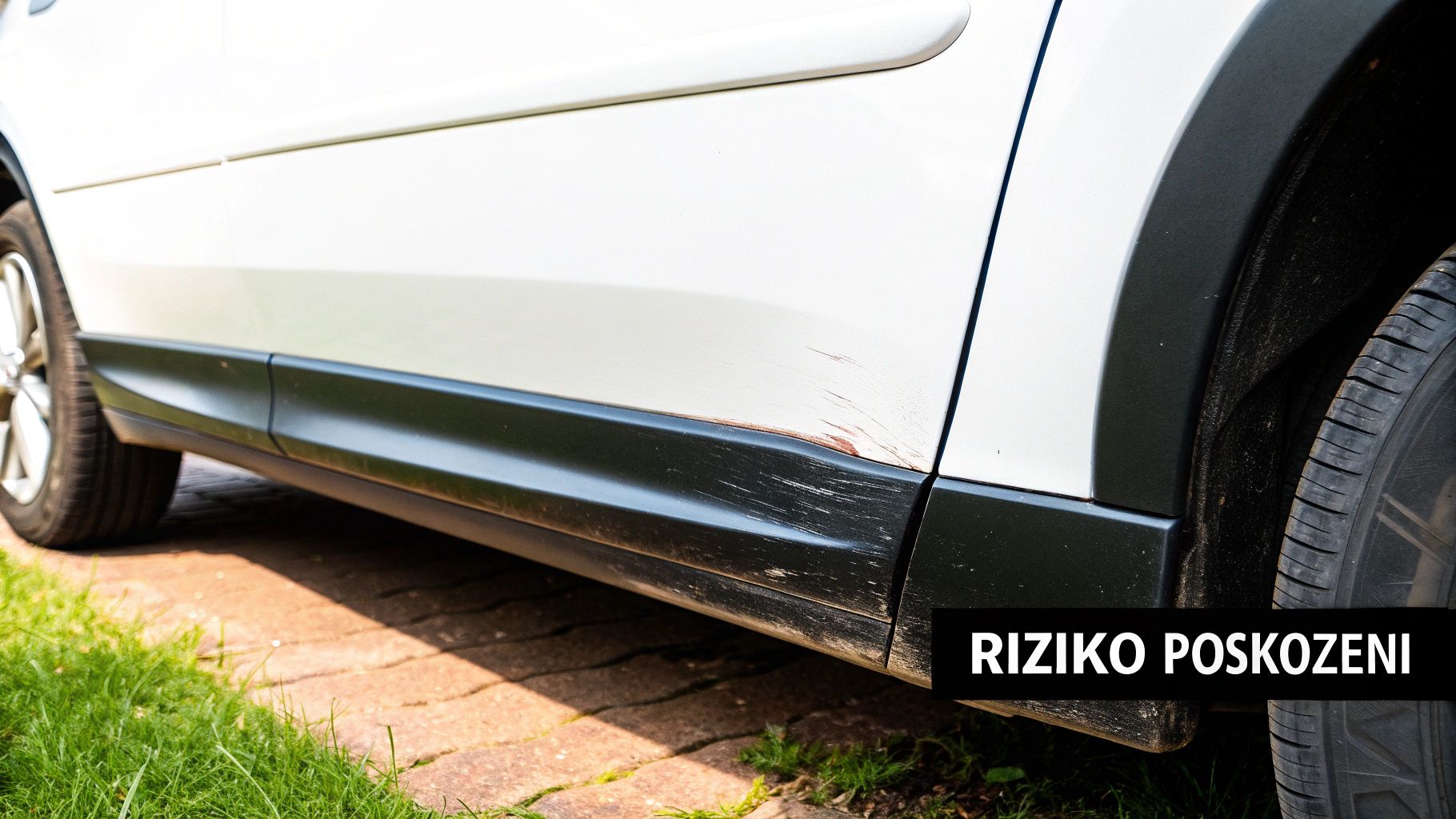 Bílý automobil se znatelnými škrábanci a rzí na boku, poblíž zadního kola, s textem RIZIKO POŠKOZENÍ.