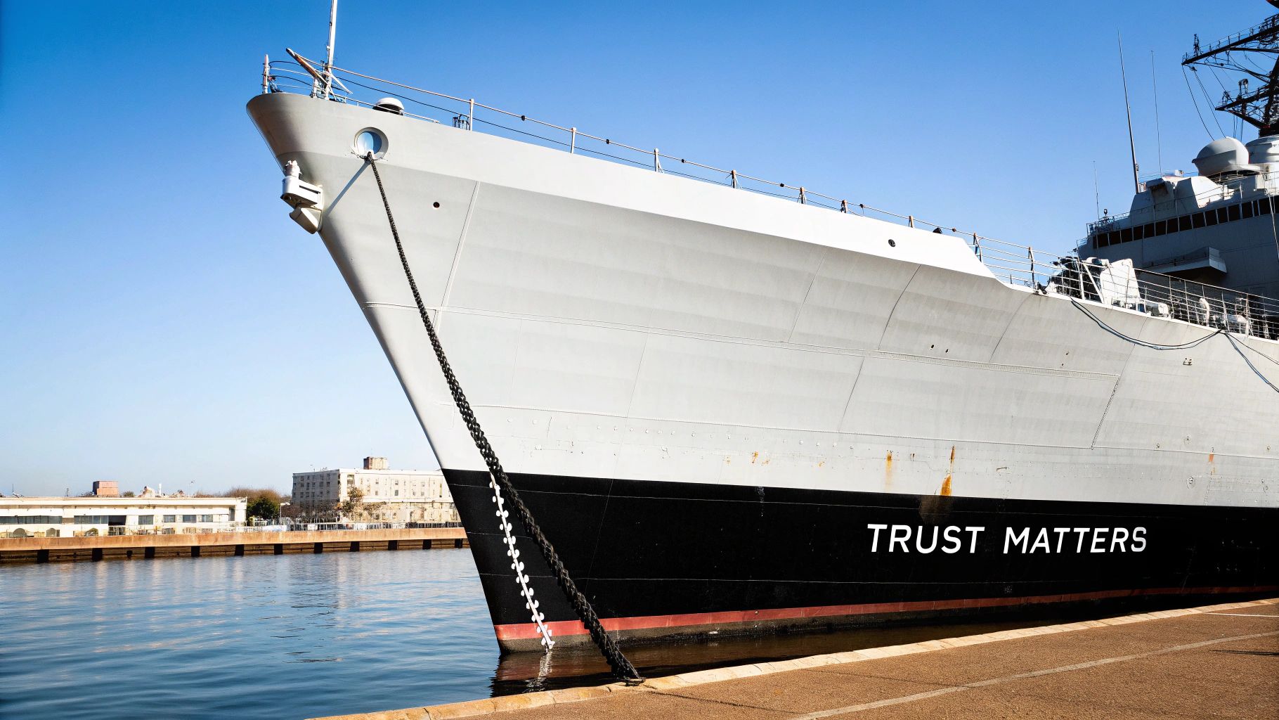Close-up of a large grey naval ship with 'TRUST MATTERS' on its hull, docked in a sunny harbor.