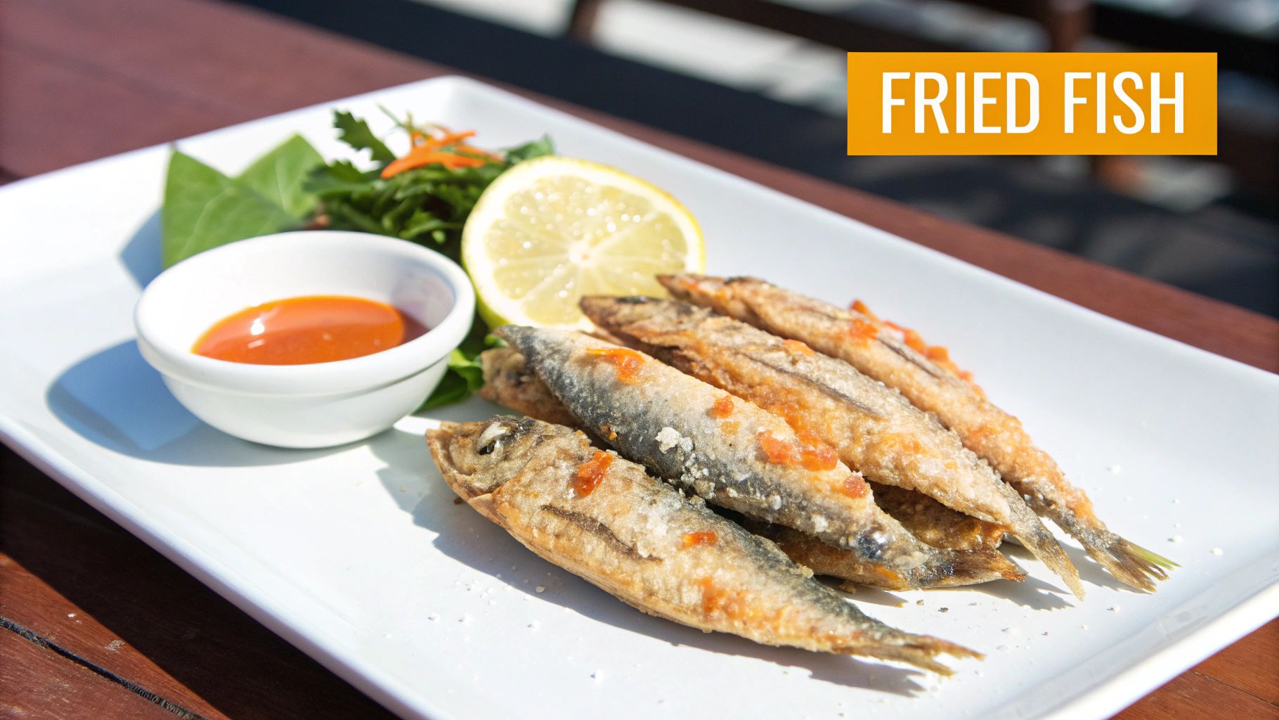 A plate of golden-brown fried fish, garnished with a lemon slice, fresh greens, and a bowl of red dipping sauce.