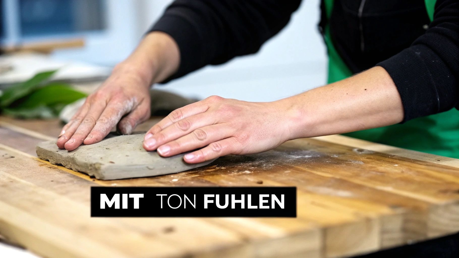 Eine Person formt mit beiden Händen Ton auf einem Holztisch, betont die taktile Erfahrung.
