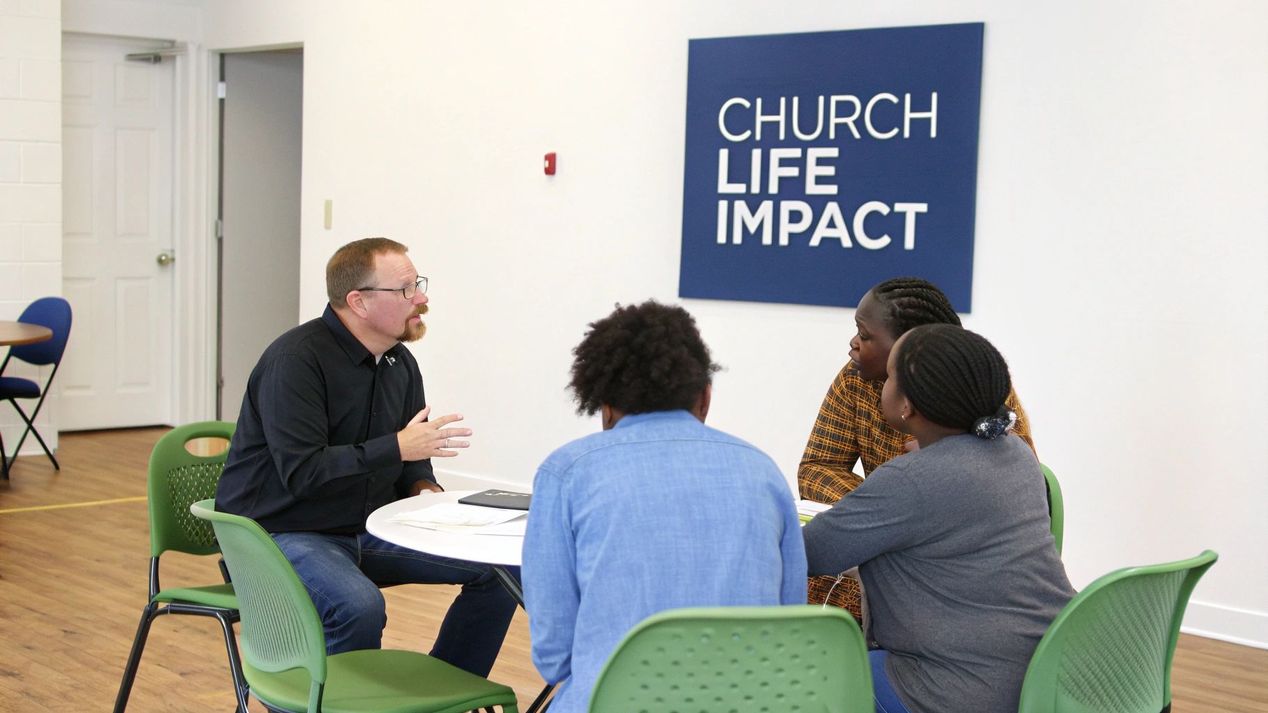 A man gestures while speaking to three women seated at a table during a church life impact discussion.