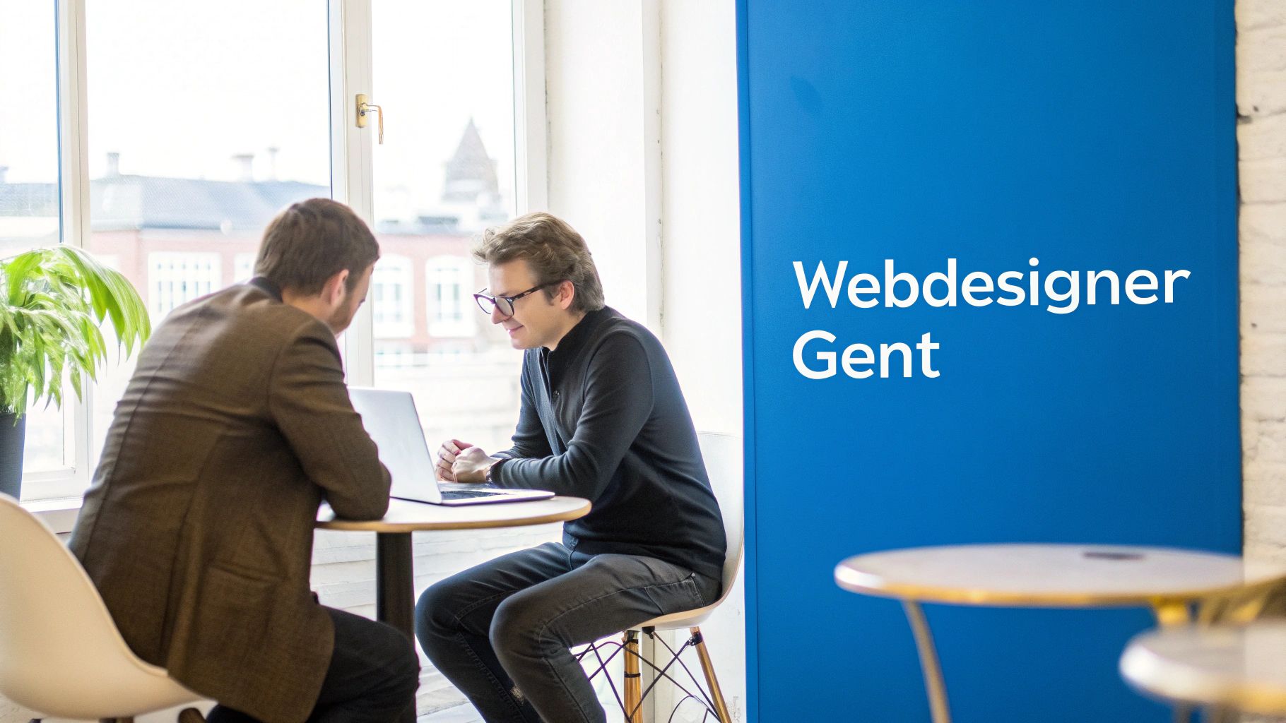 Twee mannen bespreken iets bij een laptop in een lichte ruimte, met een blauw spandoek met 'Webdesigner Gent' ernaast.