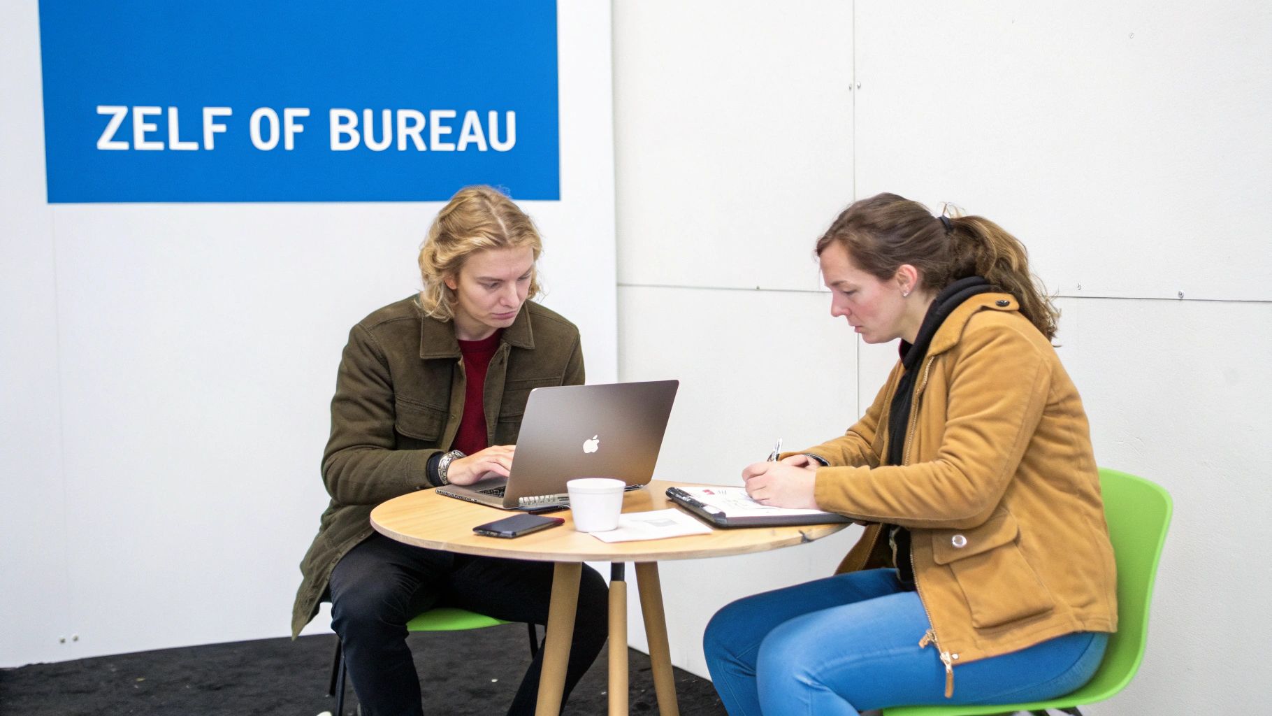 Twee mensen werken samen aan een tafel; een persoon typt op een laptop, de ander maakt aantekeningen.
