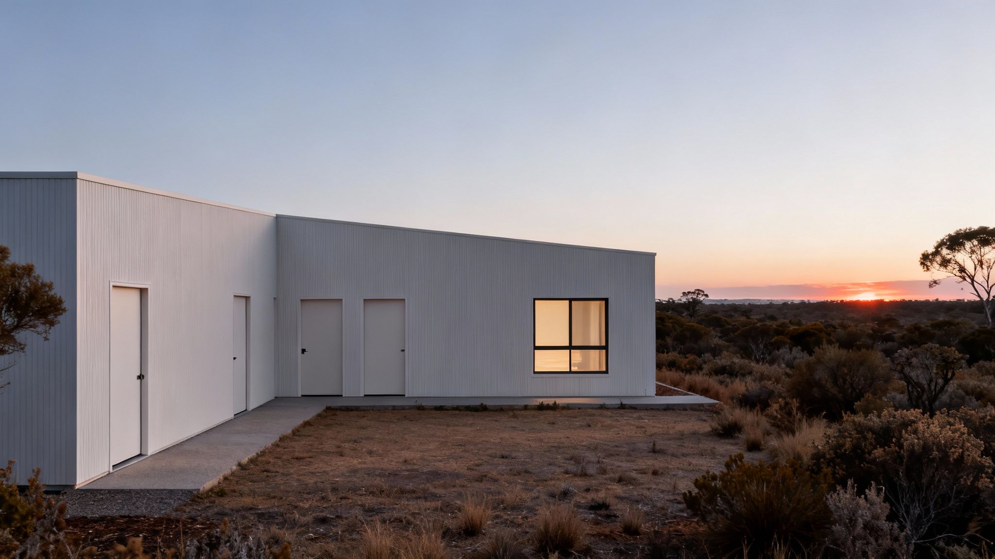 A modern white house with a single illuminated window against a sunset in a rural bushland.