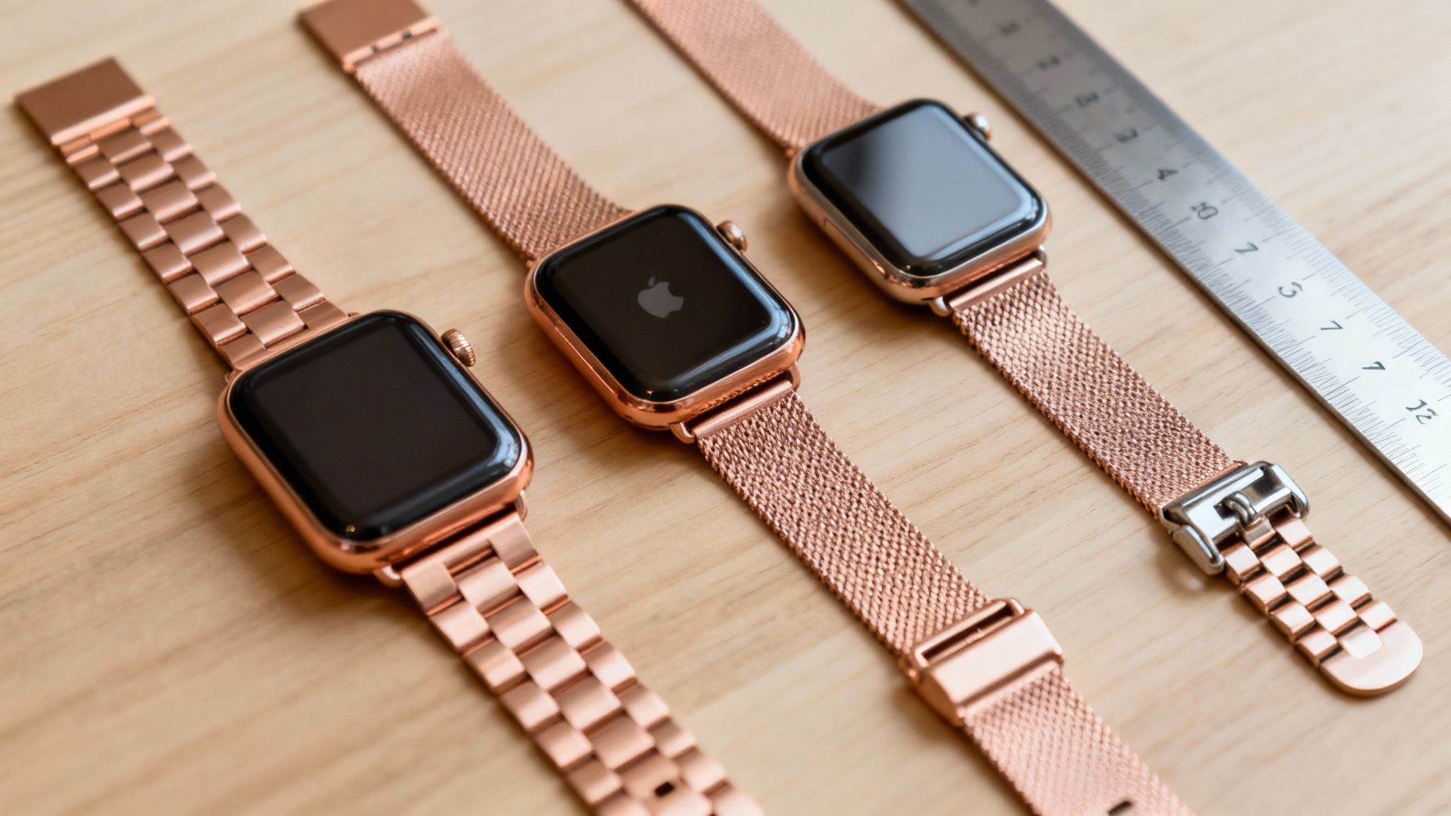 Three rose gold Apple Watches with different metal straps laid out on a light wooden surface next to a ruler.