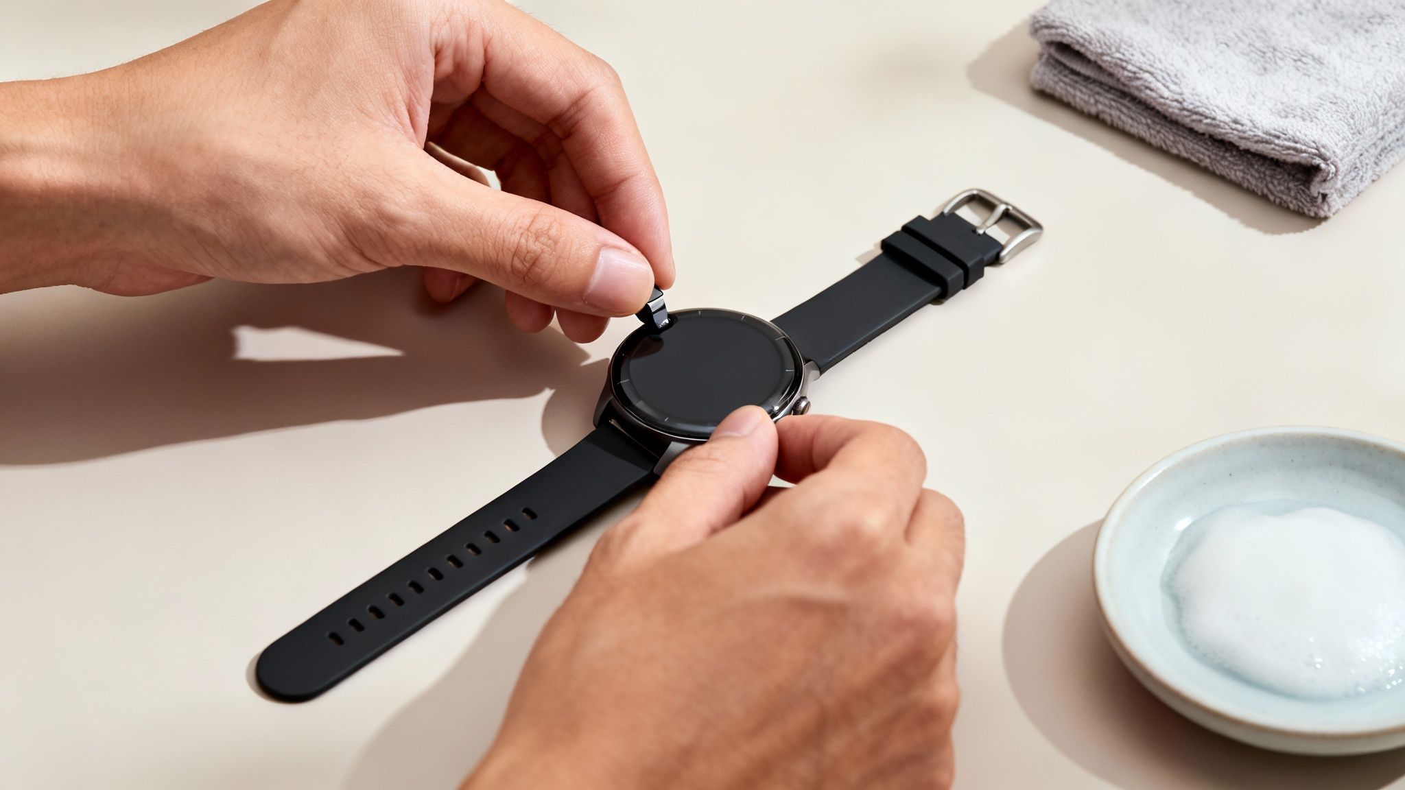 A person's hands are adjusting a black silicone strap on a round smartwatch.