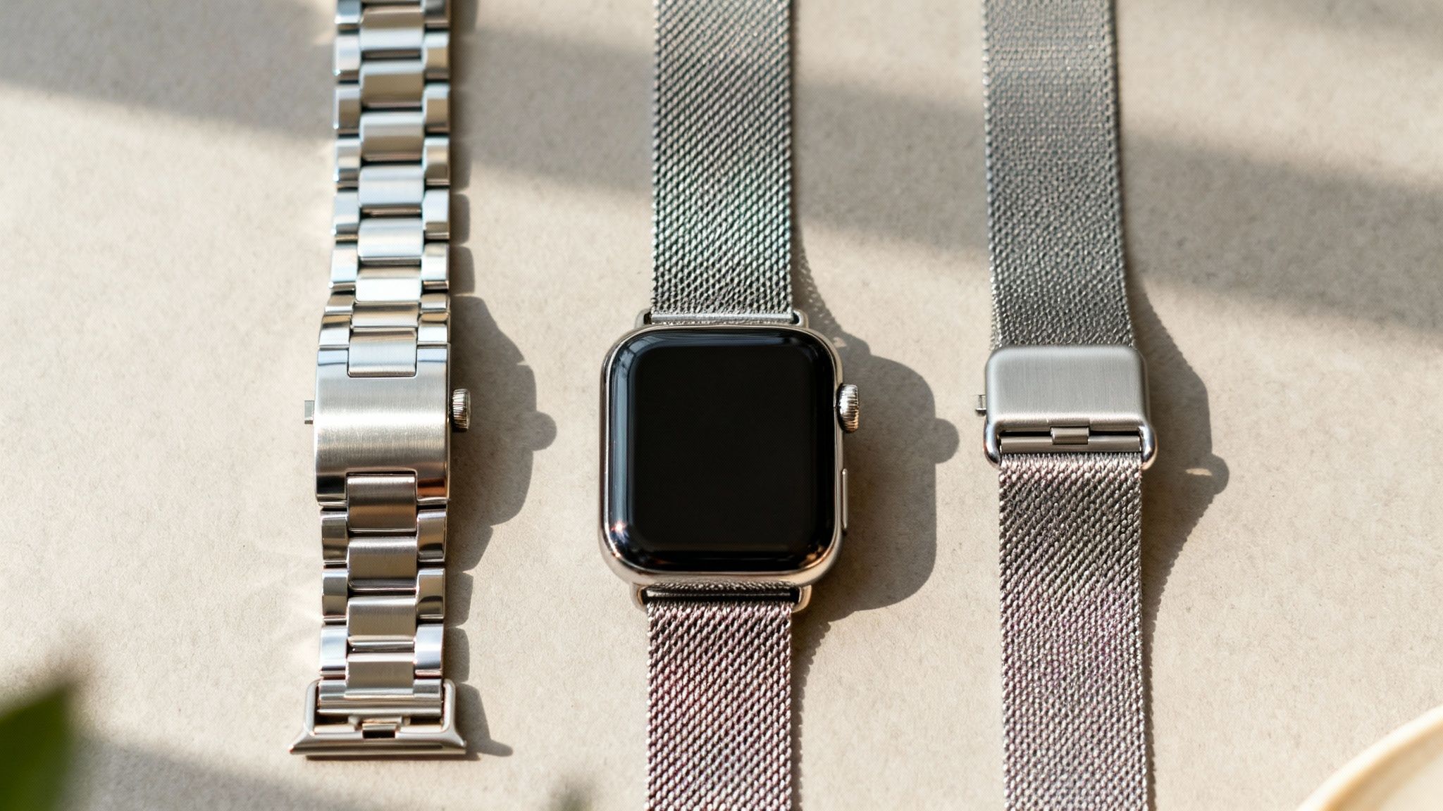 Three different silver metal watch bands for Apple Watch, one attached to a smart watch.