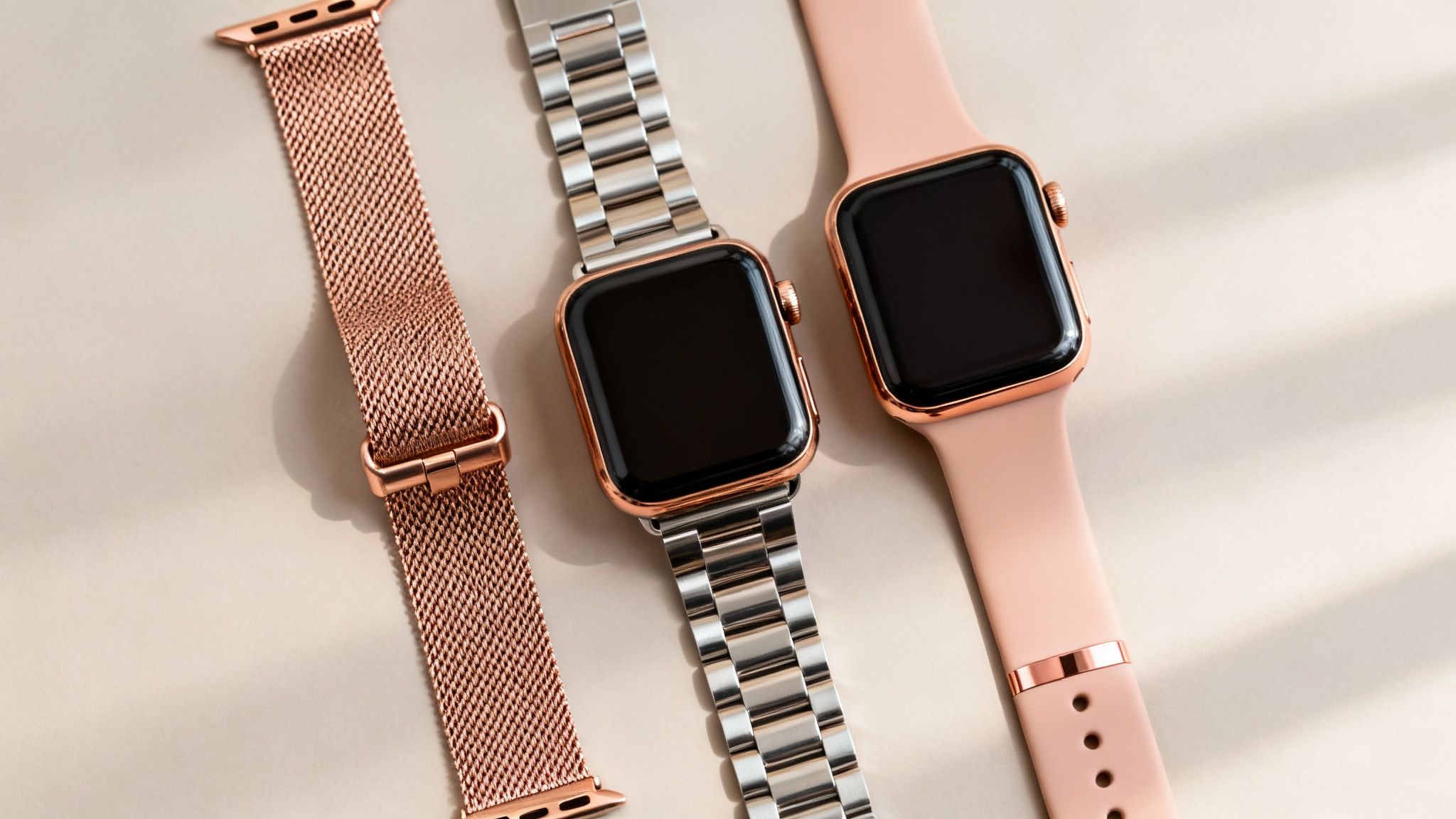 Three Apple Watch bands, including a rose gold mesh, silver link, and pink silicone, on a beige surface.