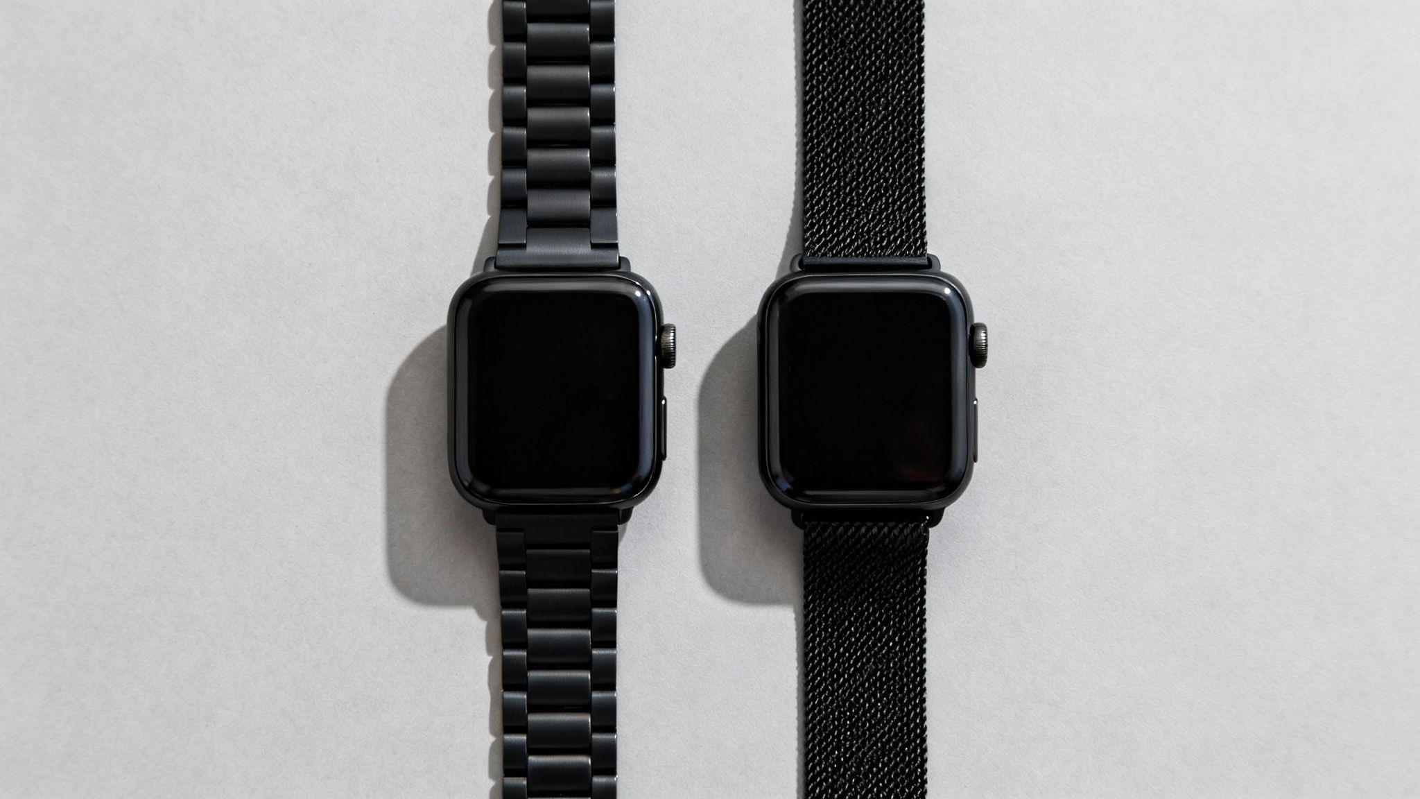 Two black smartwatches with different metal bands, one link and one mesh, on a light background.
