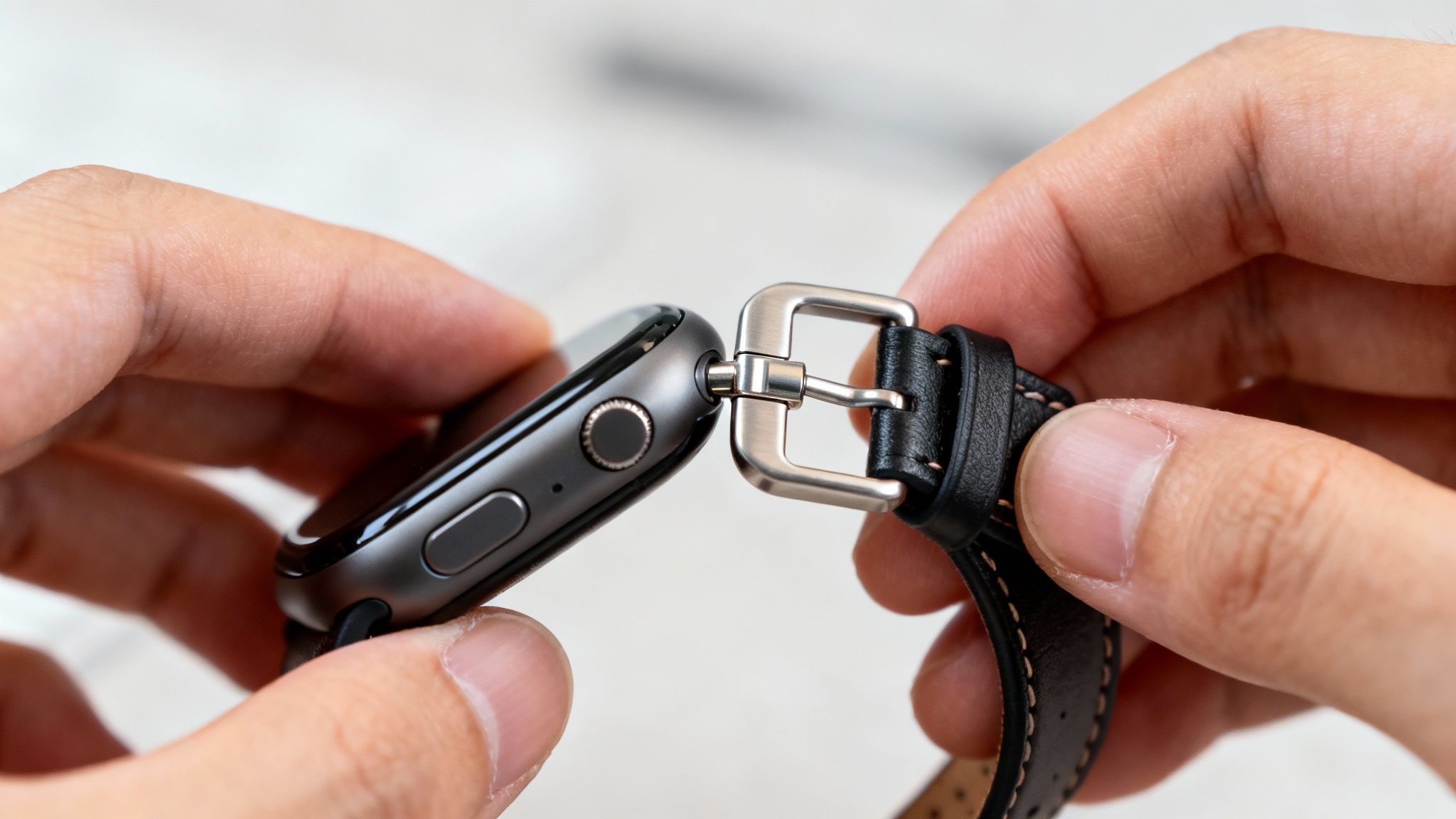 Close-up of hands attaching a black leather band with a silver buckle to a gray Apple Watch.