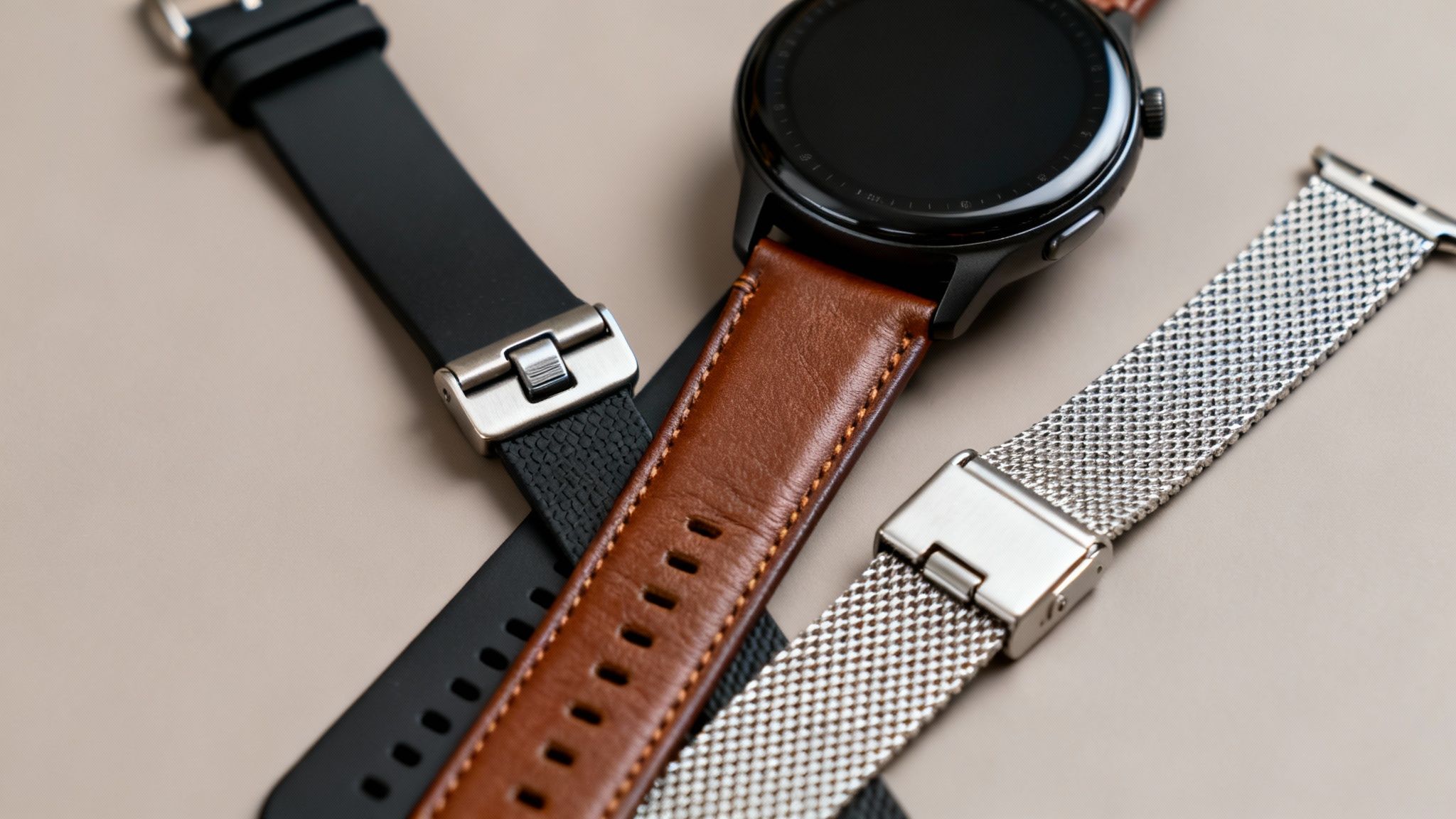 A black smartwatch with a brown leather strap, a black silicone strap, and a silver mesh strap on a beige surface.