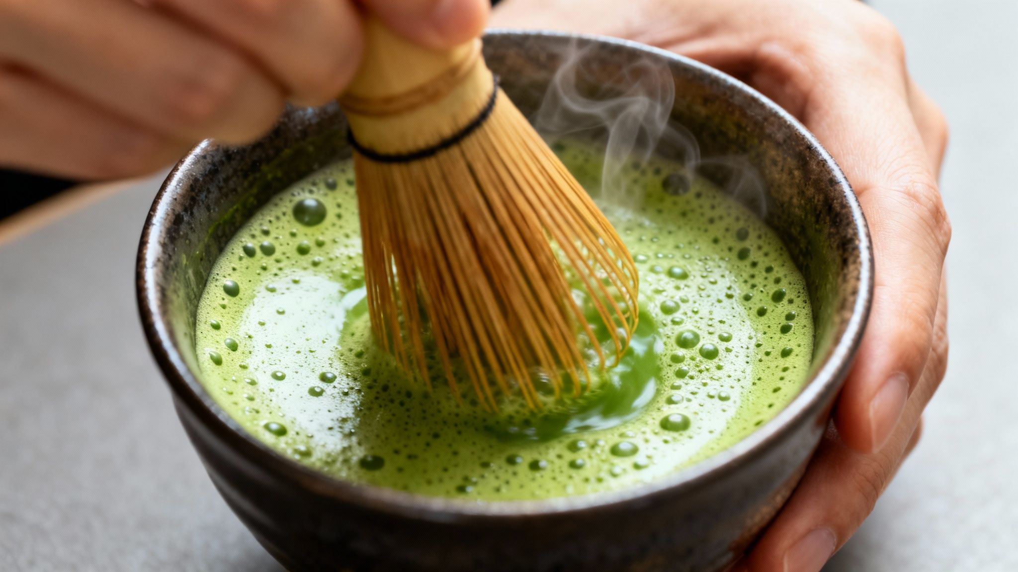 Close-up of hands whisking bright green matcha tea in a dark ceramic bowl with a bamboo whisk, releasing steam.