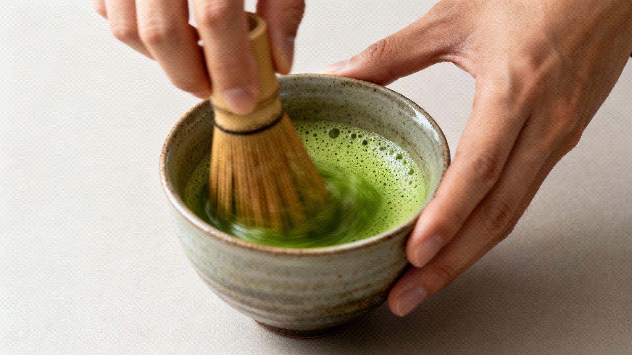 Close-up of hands whisking bright green matcha tea in a textured ceramic bowl, creating foam.