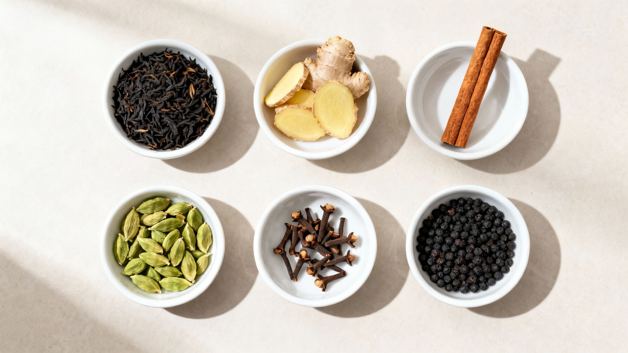 Overhead shot of six bowls with ingredients for chai tea: black tea, ginger, cinnamon, cardamom, cloves, and black peppercorns.