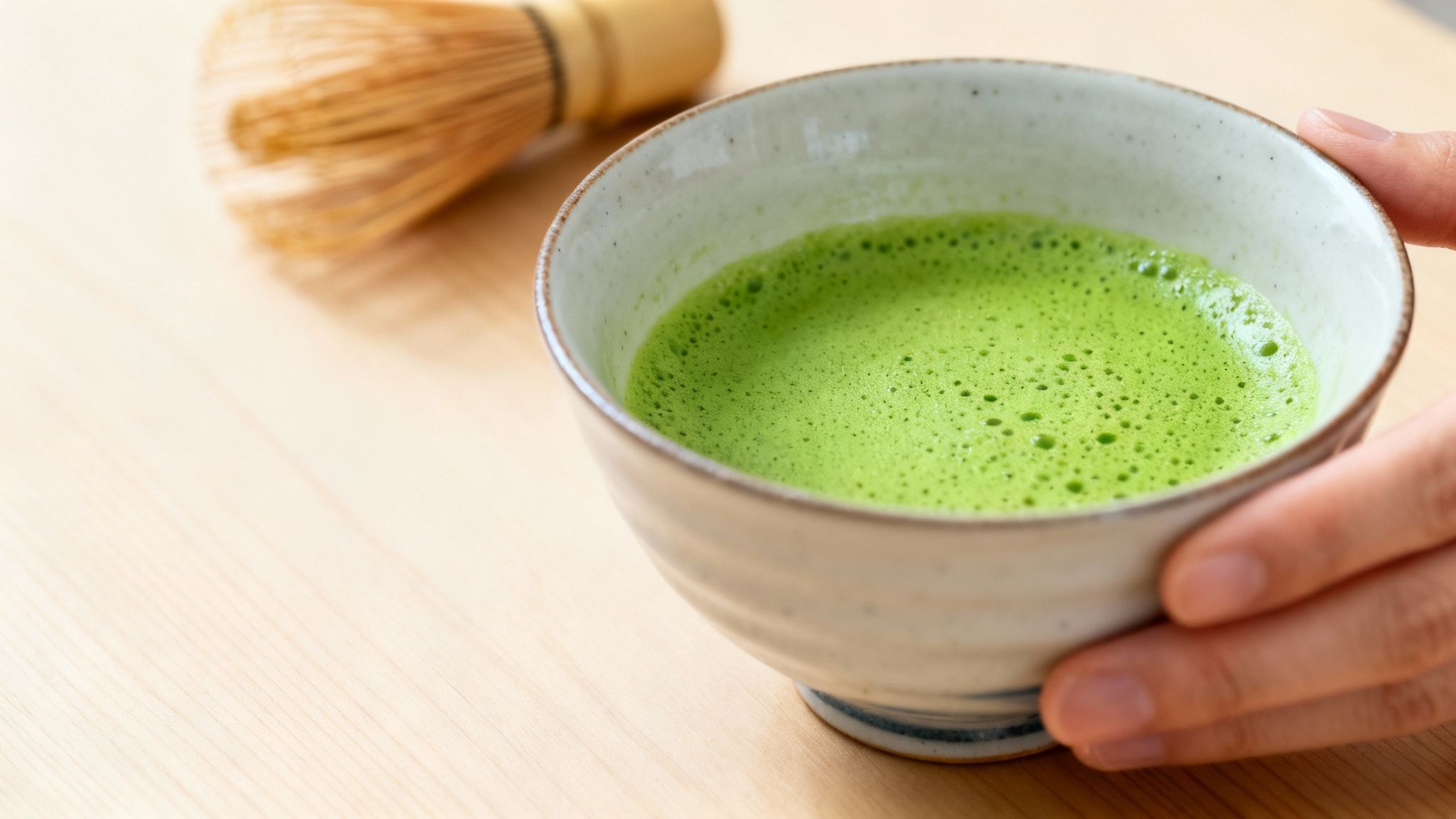 Hands gently hold a ceramic bowl of vibrant green frothy matcha tea on a wooden table.