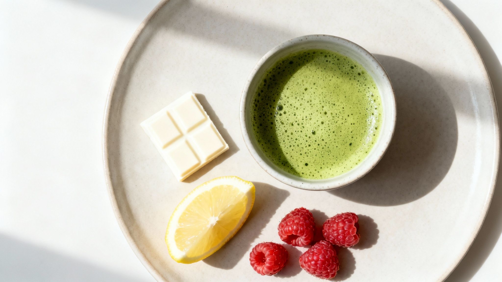 A vibrant green matcha latte, white chocolate bar, lemon wedge, and raspberries on a plate.
