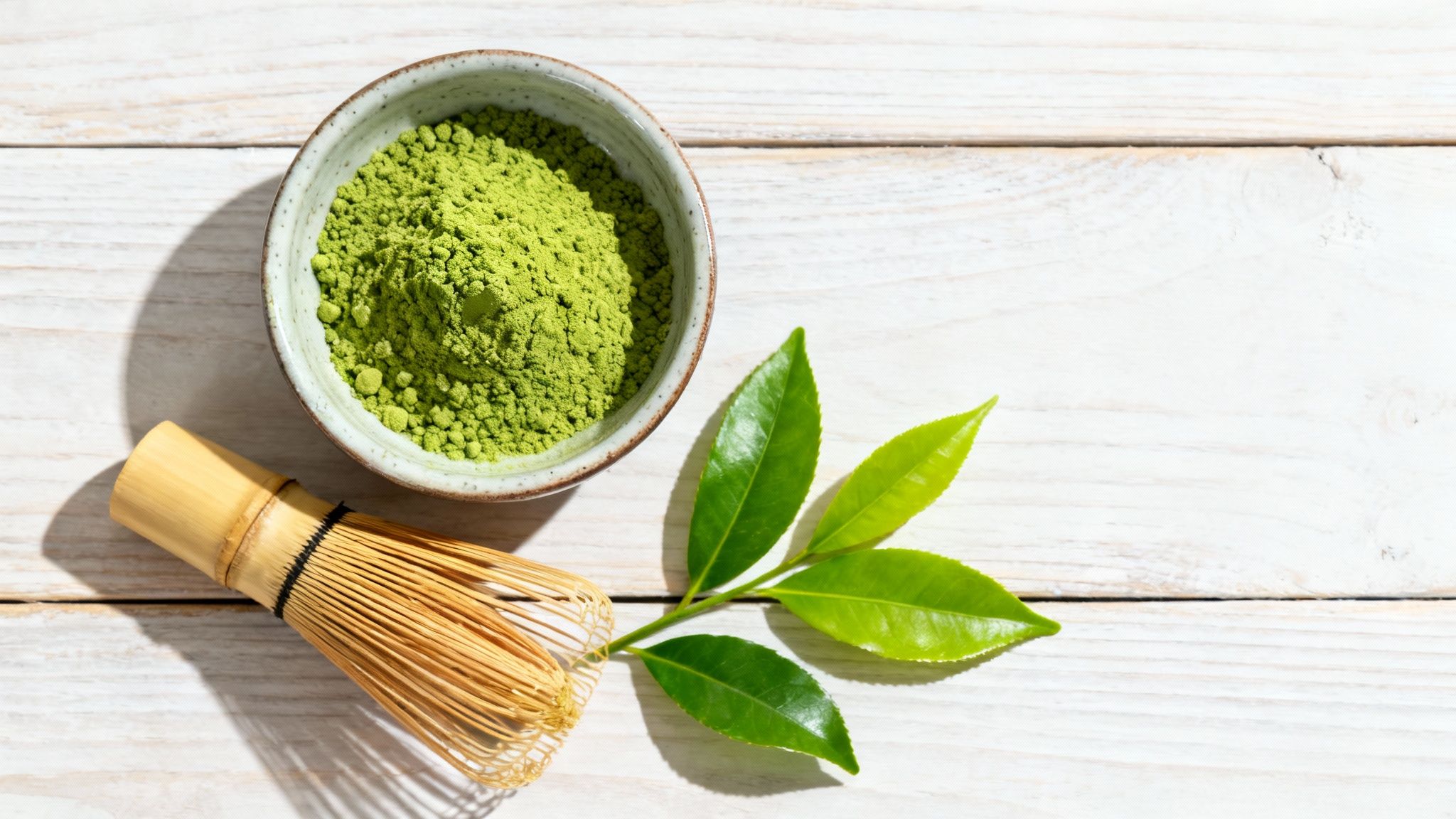 Matcha green tea powder in a bowl with a bamboo whisk and fresh tea leaves on wood.