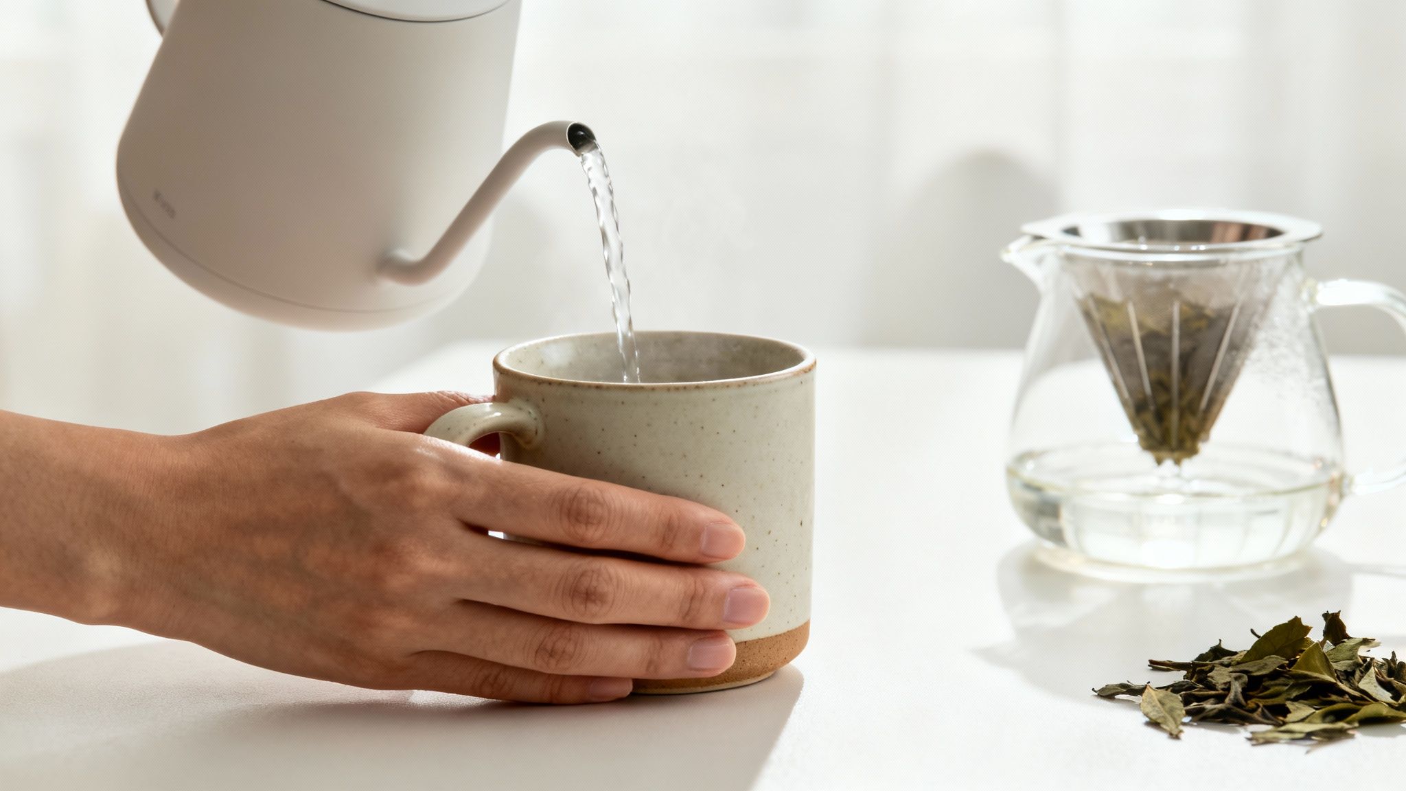 A serene scene showing a person pouring hot water from a kettle into a glass teapot with loose tea leaves inside.