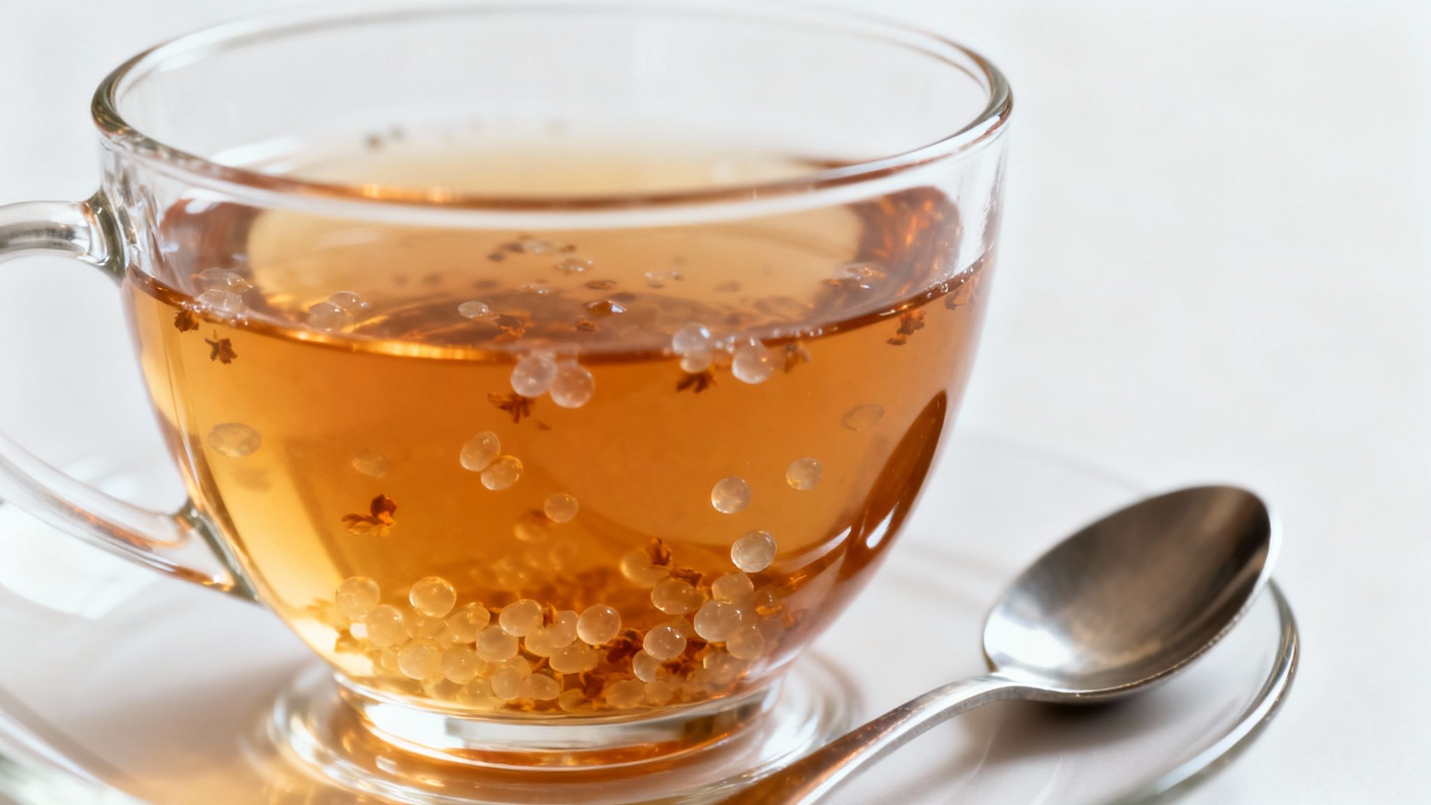 A clear glass cup of amber tea with translucent pearls and small leaves, next to a silver spoon.