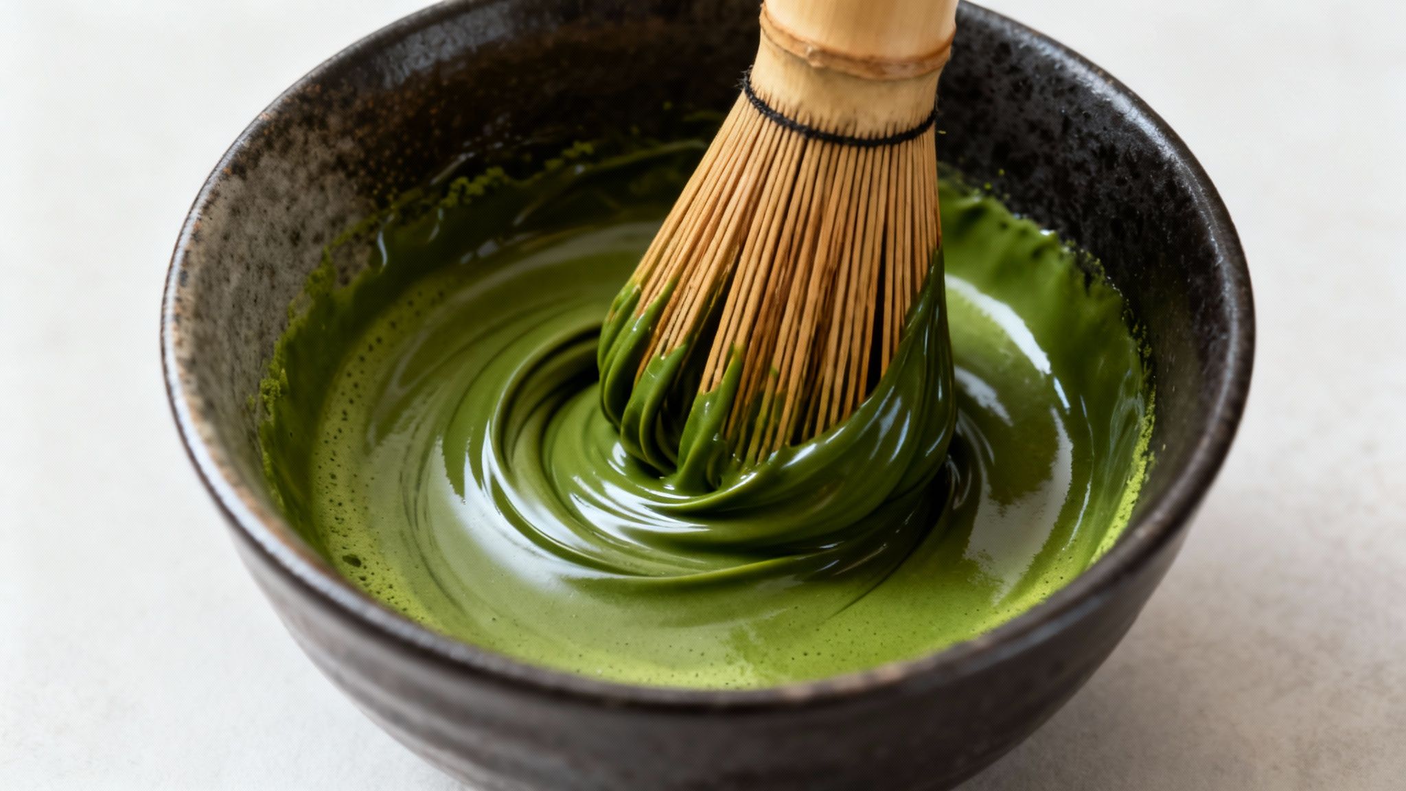 A bamboo whisk stirs vibrant green matcha tea in a dark ceramic bowl, creating a swirl.