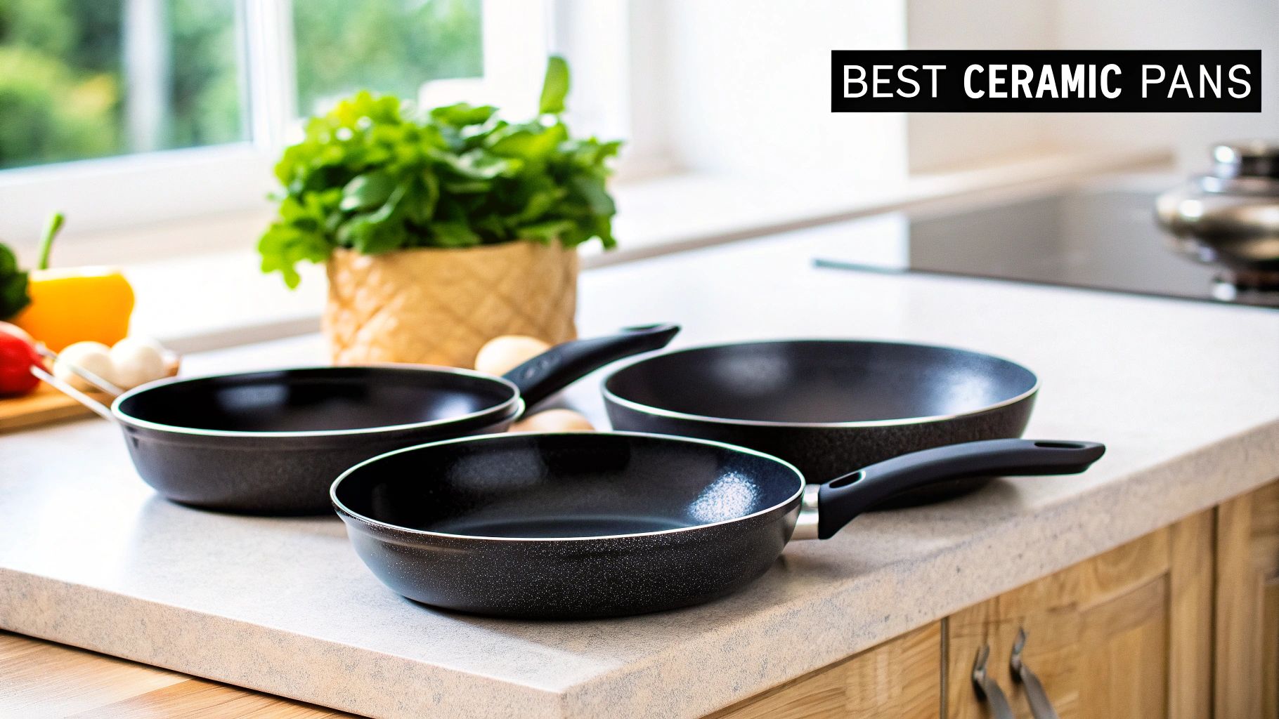 Three black ceramic frying pans sit on a modern kitchen counter with fresh vegetables and herbs.