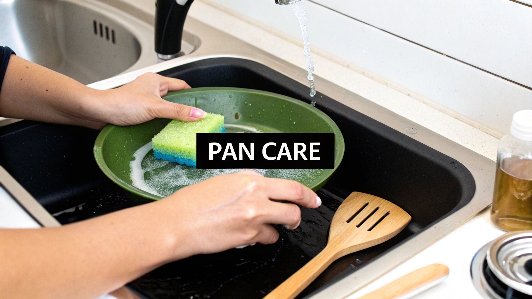 Hands washing a green pan with a sponge and soapy water in a black kitchen sink under running water.