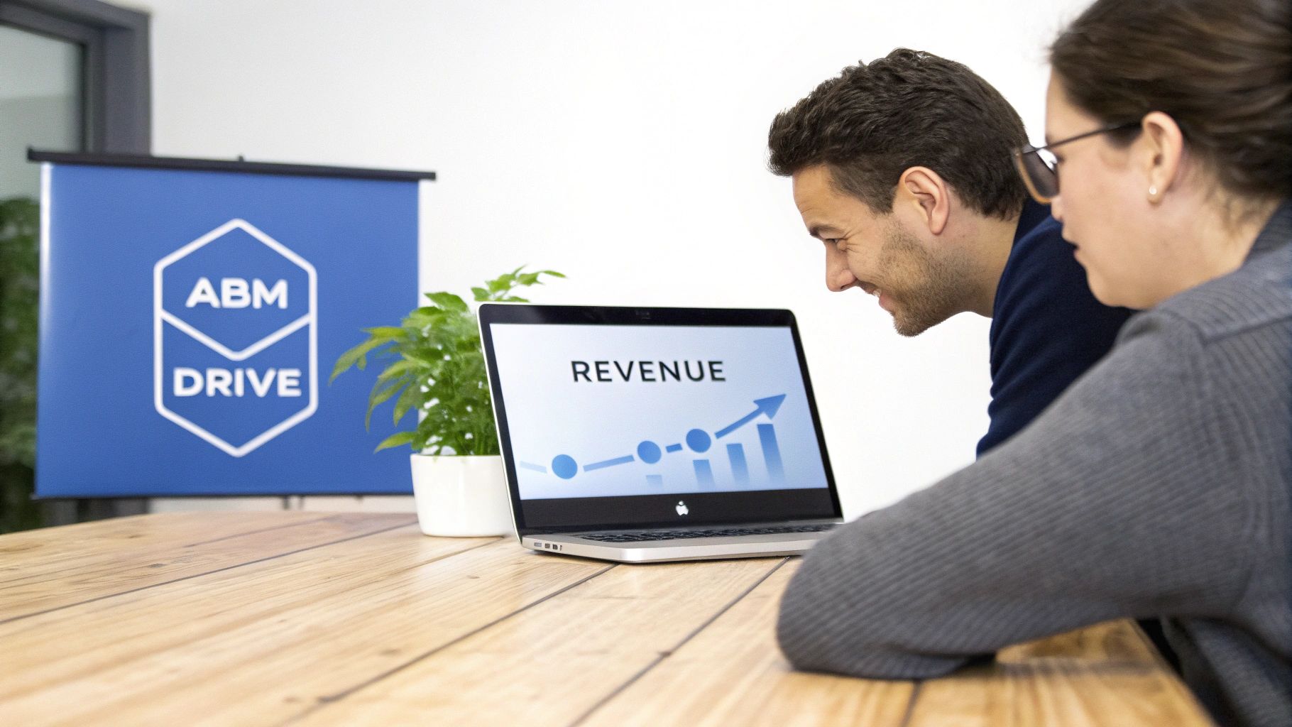 Two colleagues analyze a laptop screen displaying a rising revenue graph, with an "ABM DRIVE" banner in the background.