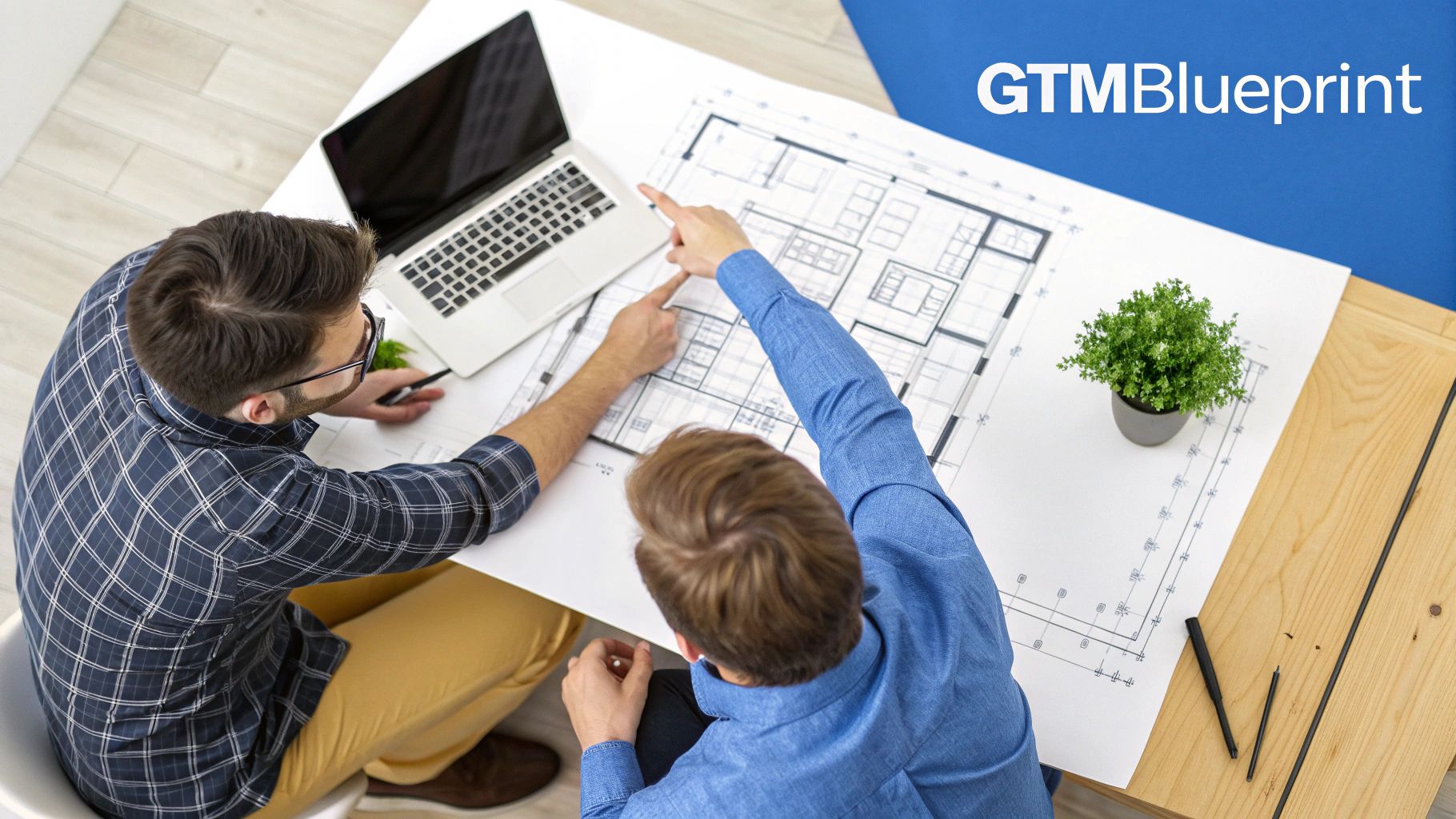Two men collaborate intently on a detailed blueprint, with a laptop and plant nearby on a table.