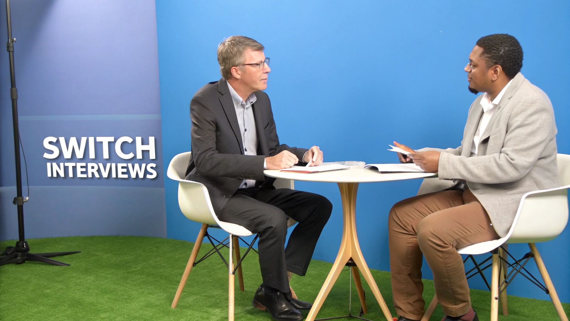 Two men are engaged in an interview at a round table, on a set with a blue wall.