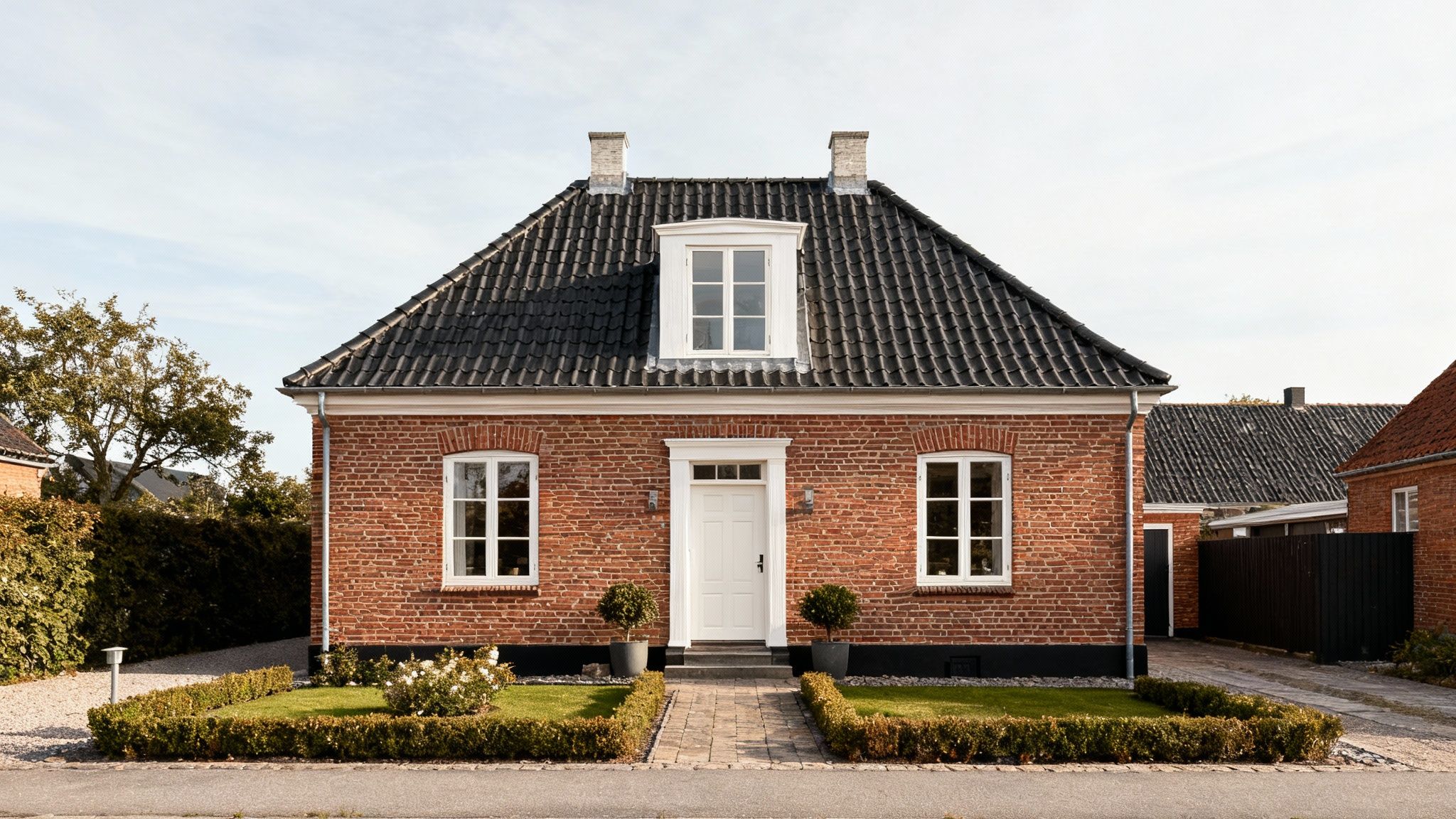 Et charmerende rødt murstenshus med sort tegltag, hvide vinduer og dør, samt en velholdt forhave.