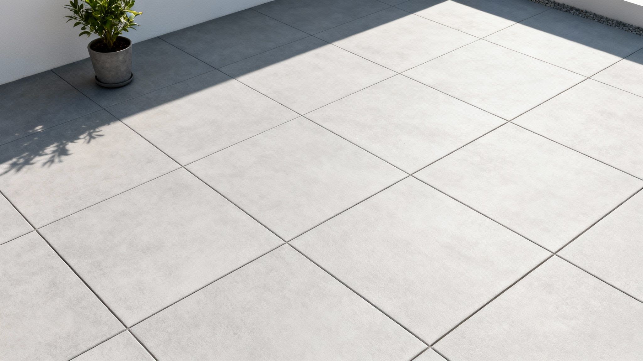 Lysegrå udendørsfliser på en terrasse med en potteplante og skygger fra solen.