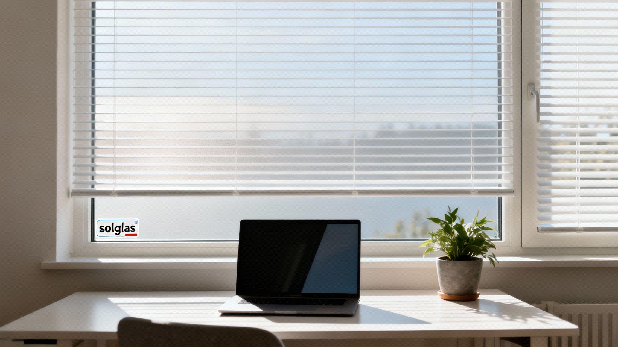 Et lyst skrivebord med en bærbar computer, en plante og hvide persienner i vinduet med et solglas-mærkat.
