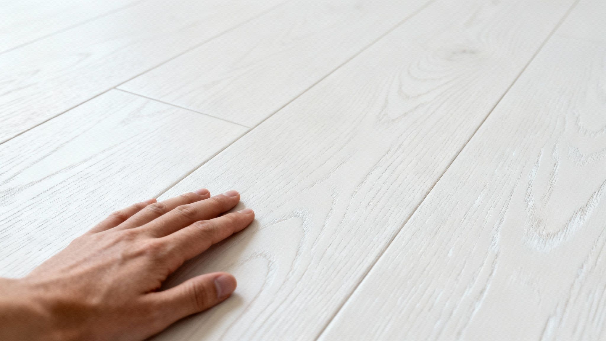 Nærbillede af en hånd, der rører ved et lyst hvidt trægulv med mat lakfinish.