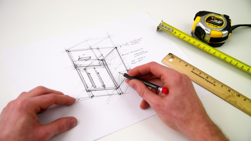 Hænder tegner et møbeldesign på papir med blyant, målebånd og lineal på et hvidt bord.