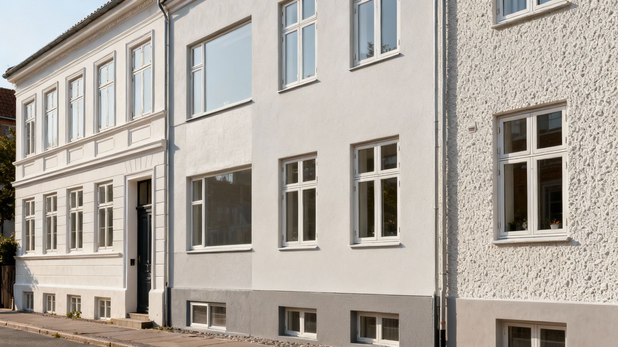Tre hvide og lysegrå bygningsfacader med vinduer, beliggende ved en gade i solskin.