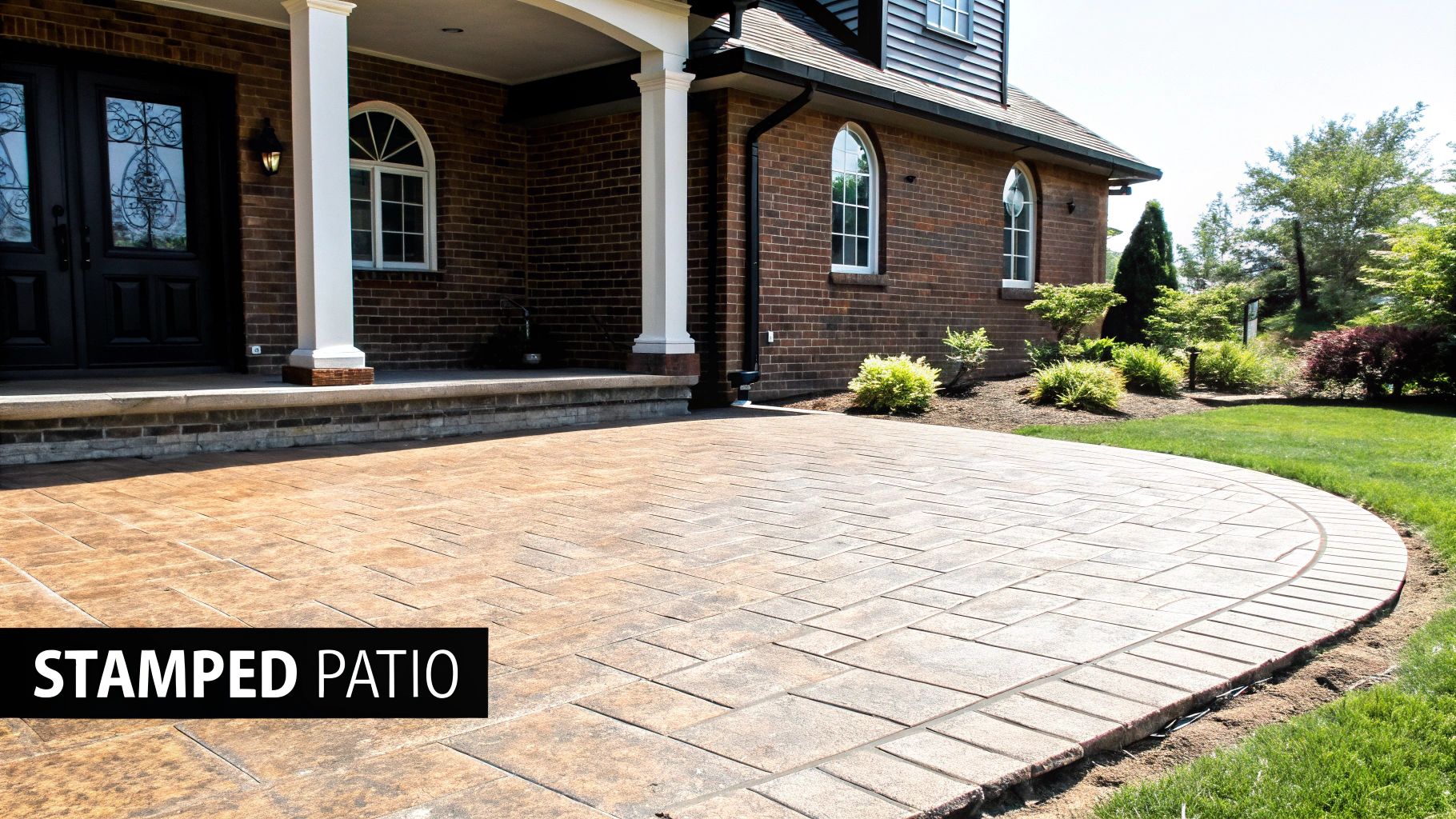 A beautiful brown stamped concrete patio curves around a brick house with green landscaping.