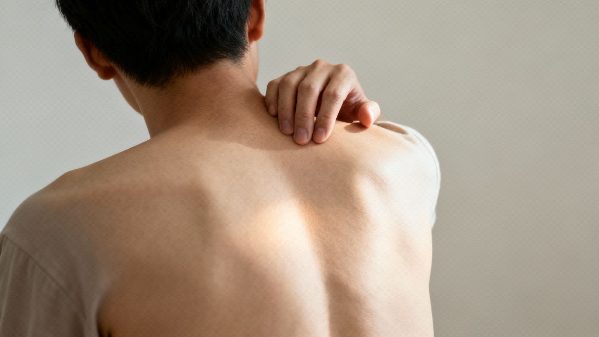 Person reaching back with hand to scratch upper shoulder blade area indicating discomfort
