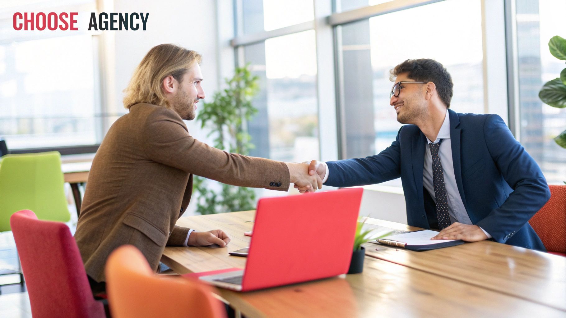 Two businessmen shaking hands over a table with a laptop on it 