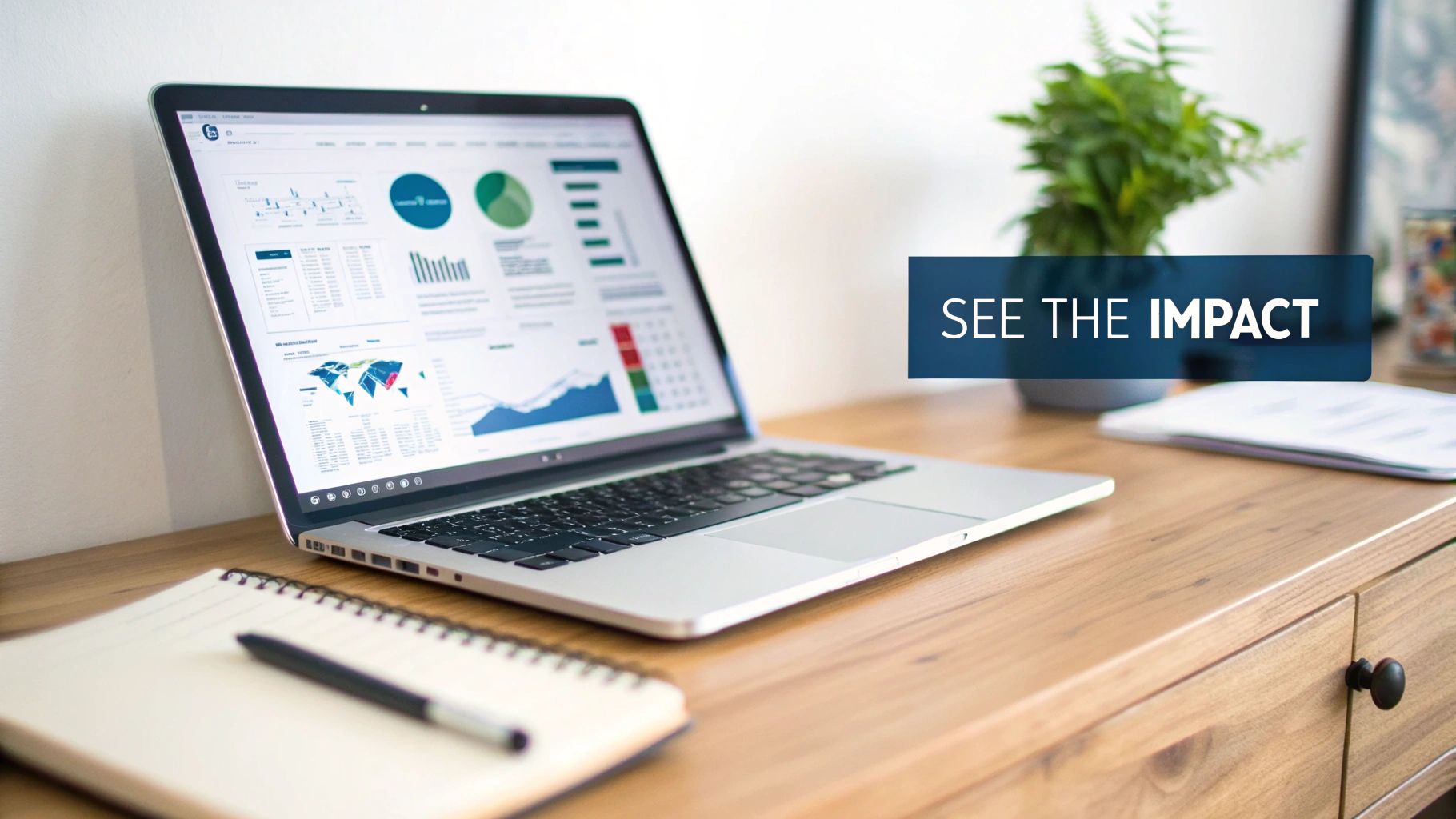 A laptop on a wooden desk displays data dashboards with charts and graphs, next to a notebook and plant.