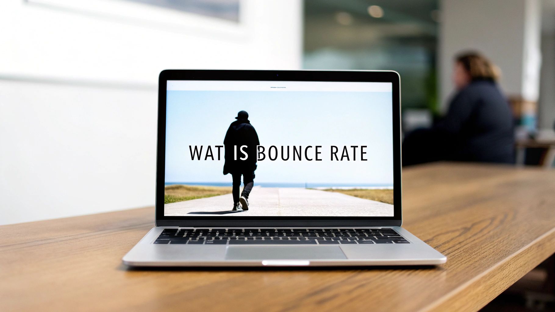 Een laptop op een houten bureau met de tekst 'WAT IS BOUNCE RATE' op het scherm, naast een persoon die wegloopt.