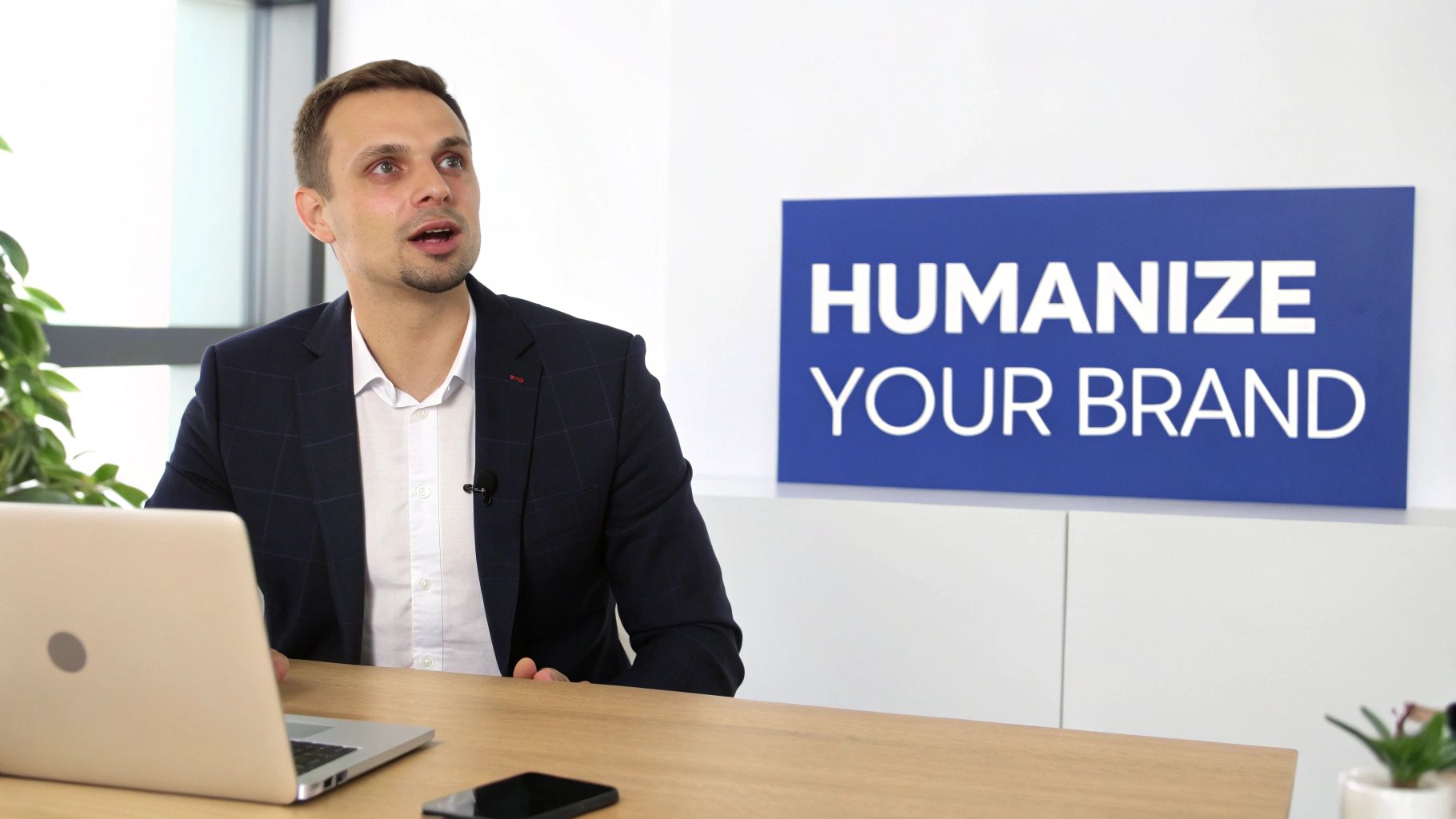 A professional man in a suit speaking at a desk with a laptop, next to a 'Humanize Your Brand' sign.