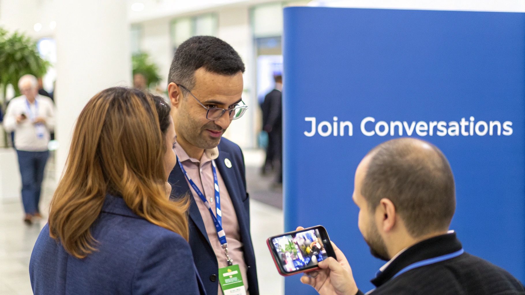 Attendees at a professional event engaging, one showing content on a smartphone near a blue banner.